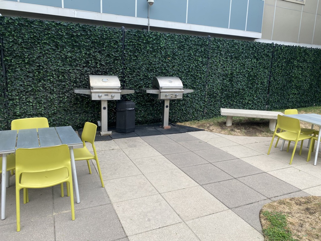Outdoor patio with two stainless steel grills, yellow chairs, a table, and a bench. Green hedge background and tiled flooring. No people present.