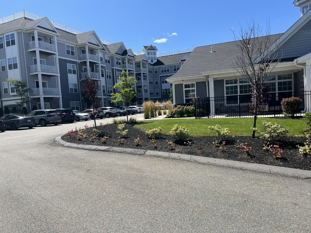 Modern residential complex with landscaped gardens, parked cars, and clear blue sky. Apartments feature balconies and pitched roofs, creating a clean, suburban atmosphere.