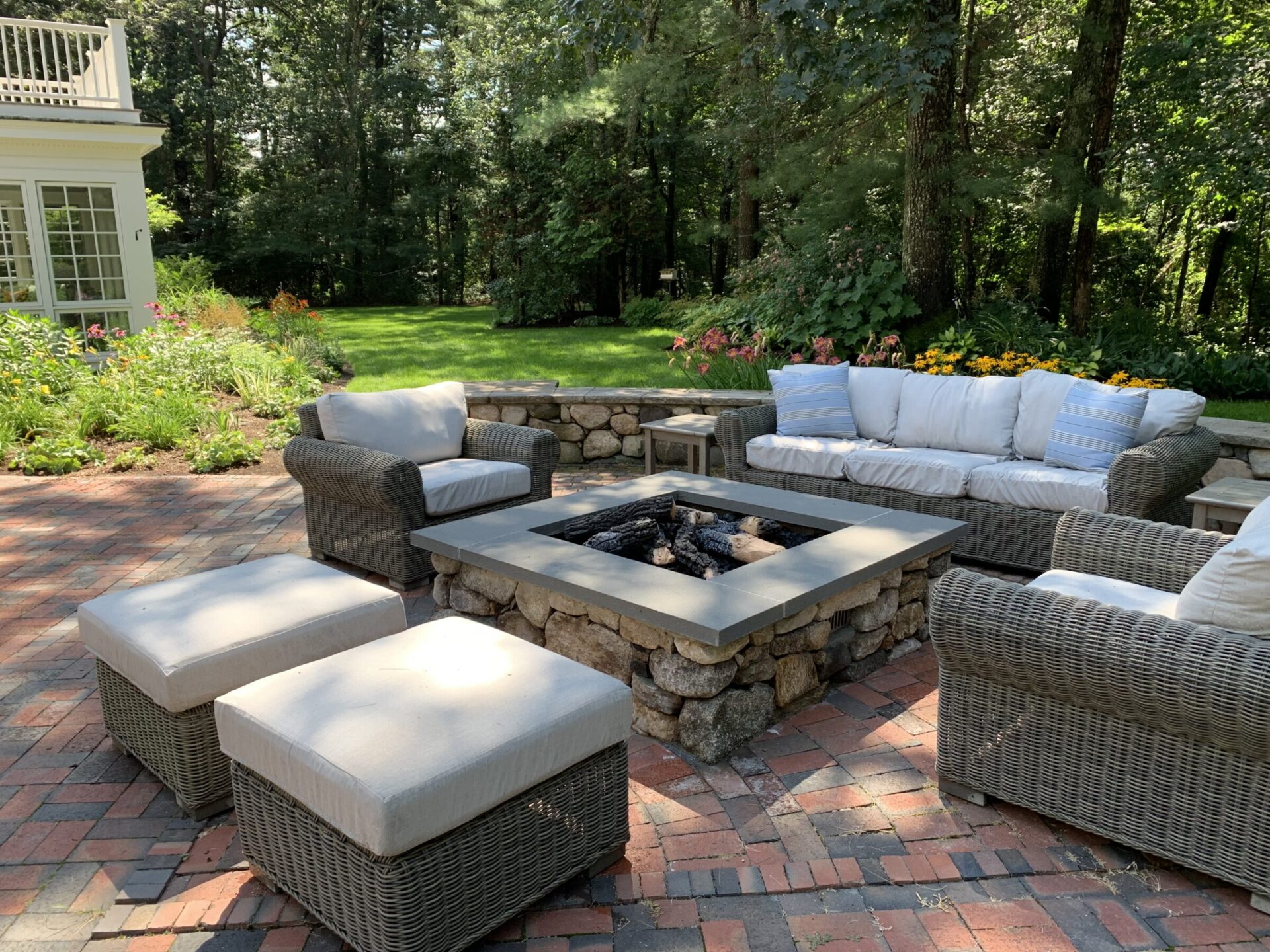Outdoor patio with wicker furniture around a stone fire pit, surrounded by lush greenery and vibrant flowers on brick paving.