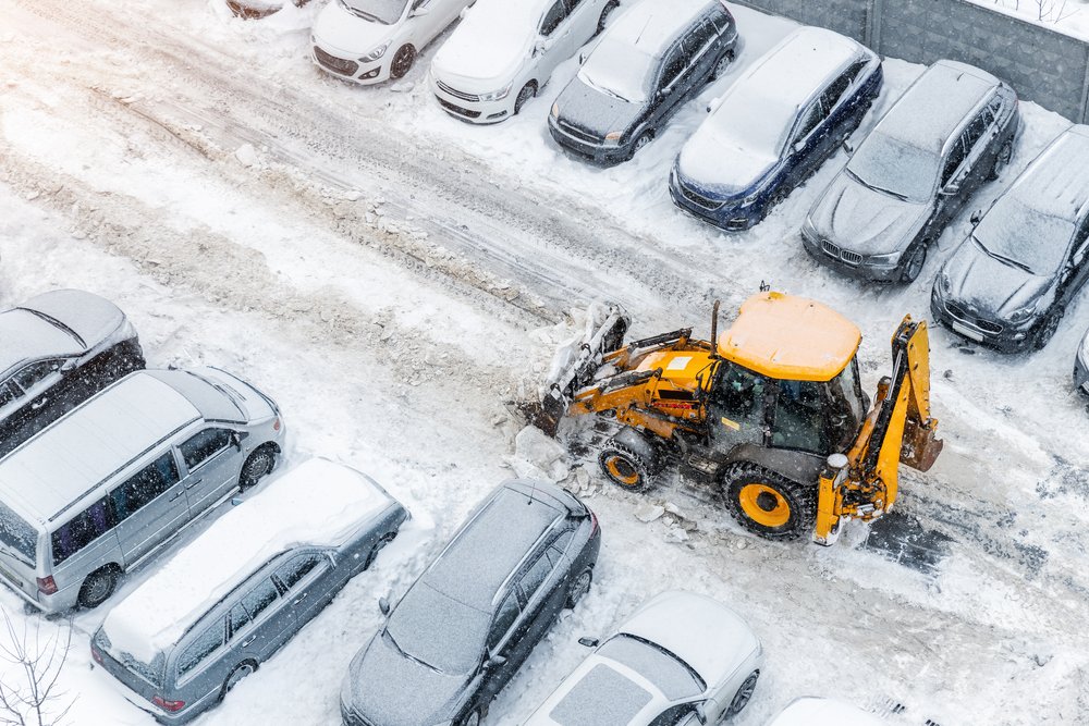 A yellow snowplow clears snow from a parking lot filled with cars, surrounded by snow-covered vehicles under winter conditions.