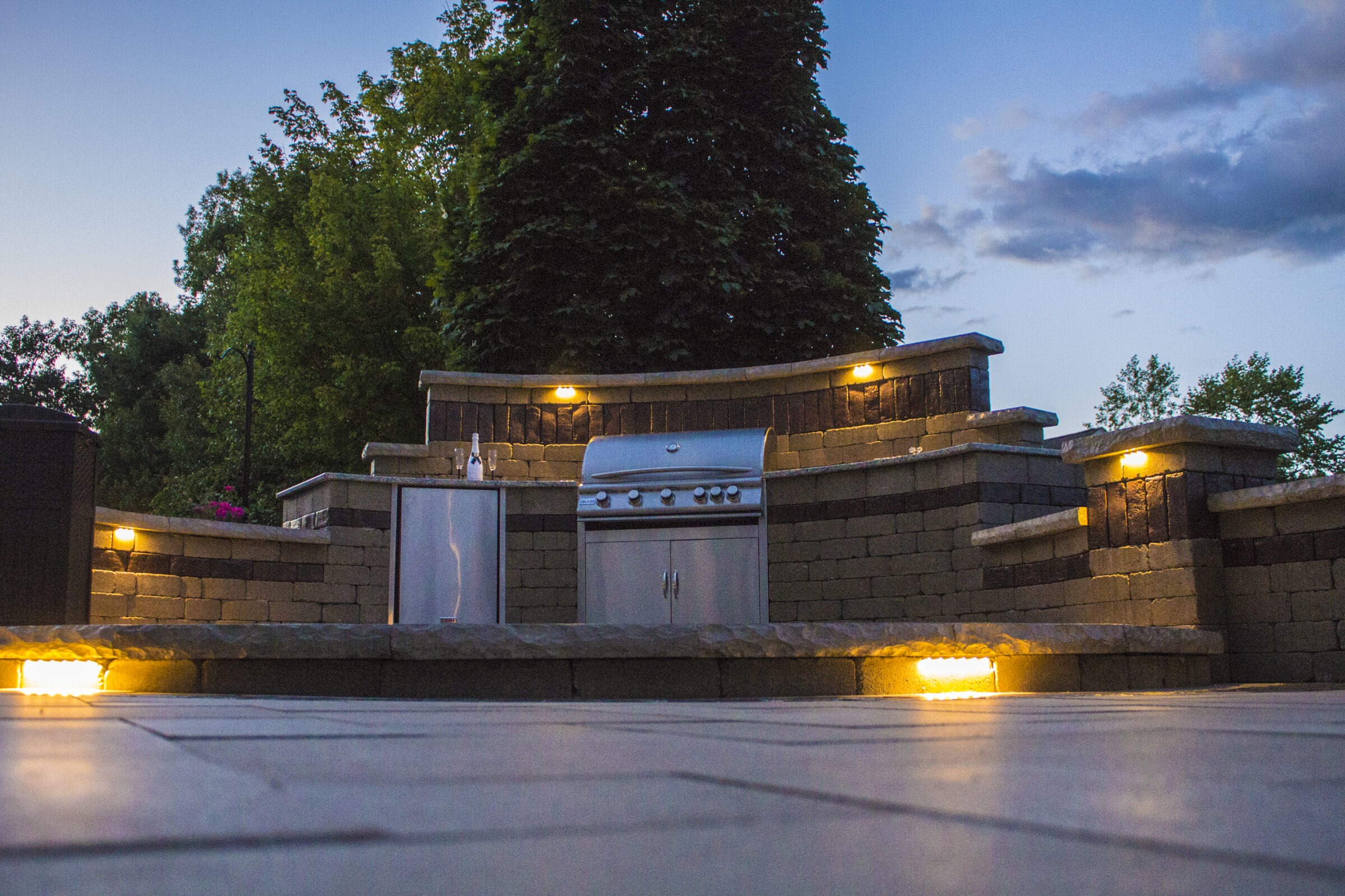 Outdoor kitchen with built-in grill, stone walls, and ambient lighting. Surrounded by trees at dusk, creating a cozy evening atmosphere.