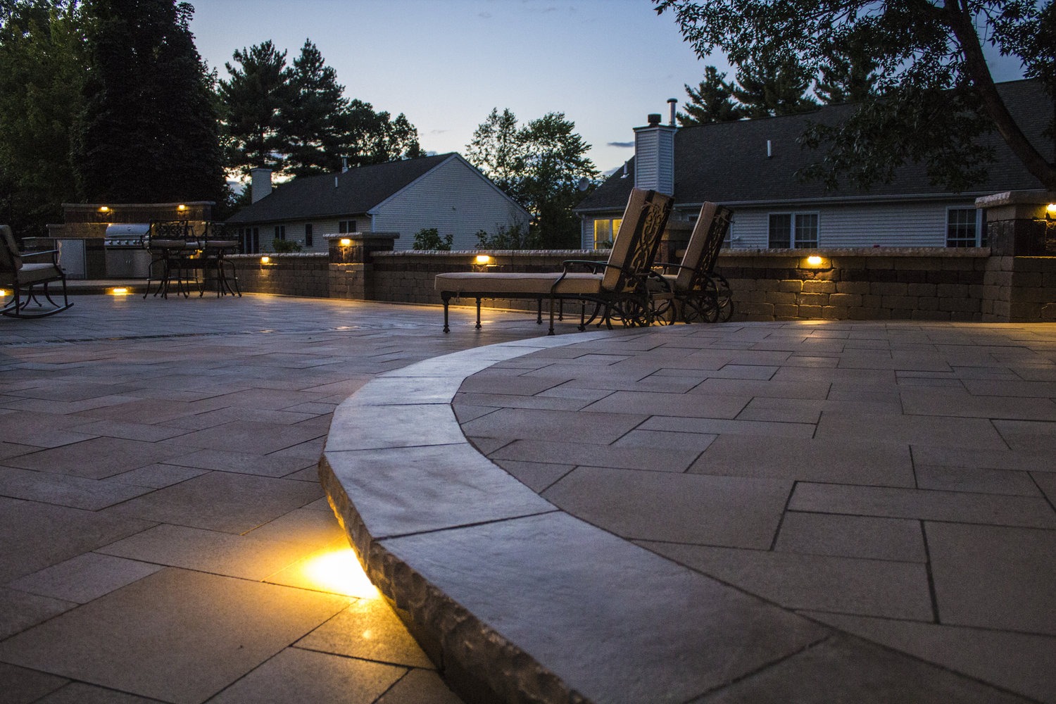 A spacious, paved patio with soft lights, lounge chairs, and trees, situated near residential houses, at dusk.
