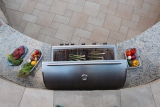 Top-down view of a grill surrounded by vegetables and meat on a curved countertop, set on a patterned tiled floor. No people visible.