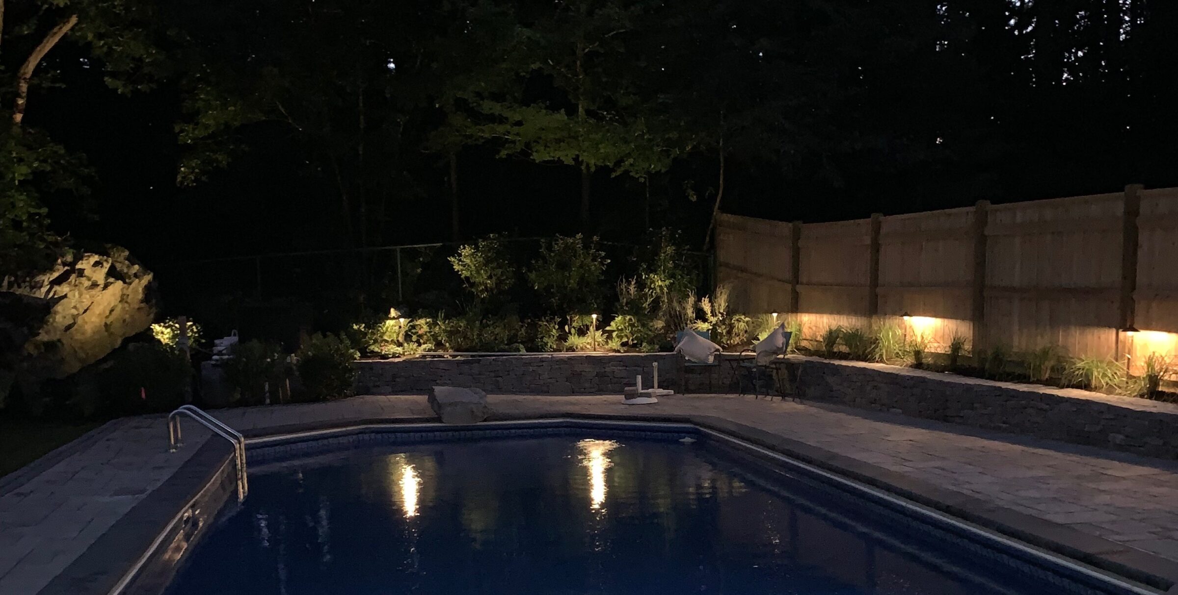 A dimly lit backyard features a serene swimming pool, surrounded by chairs and a wooden fence, amidst lush greenery under a night sky.