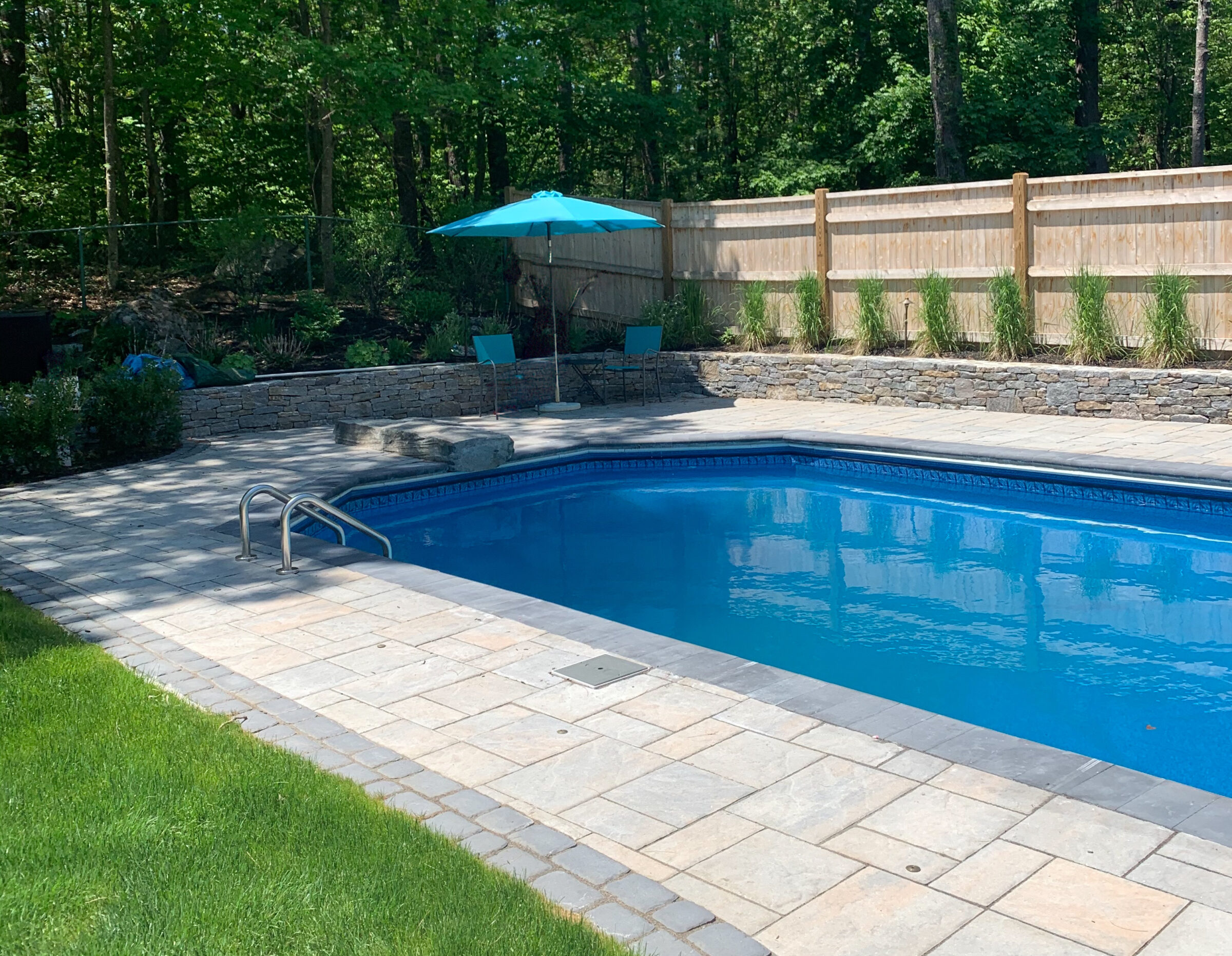 A backyard swimming pool with stone patio, surrounded by lush greenery and wooden fence, featuring chairs and a blue umbrella.