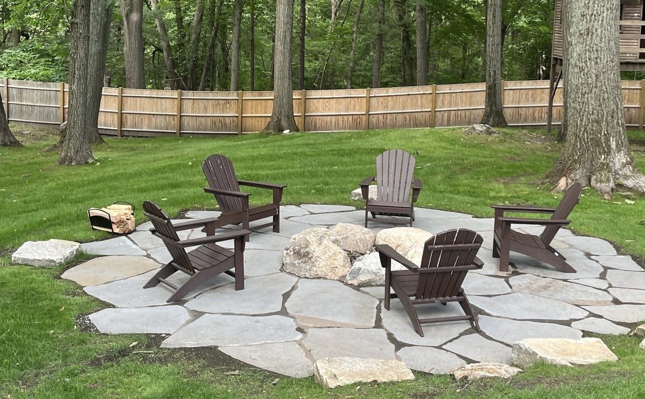 A circular stone patio features five wooden chairs and a fire pit, surrounded by trees and a wooden fence in a tranquil setting.
