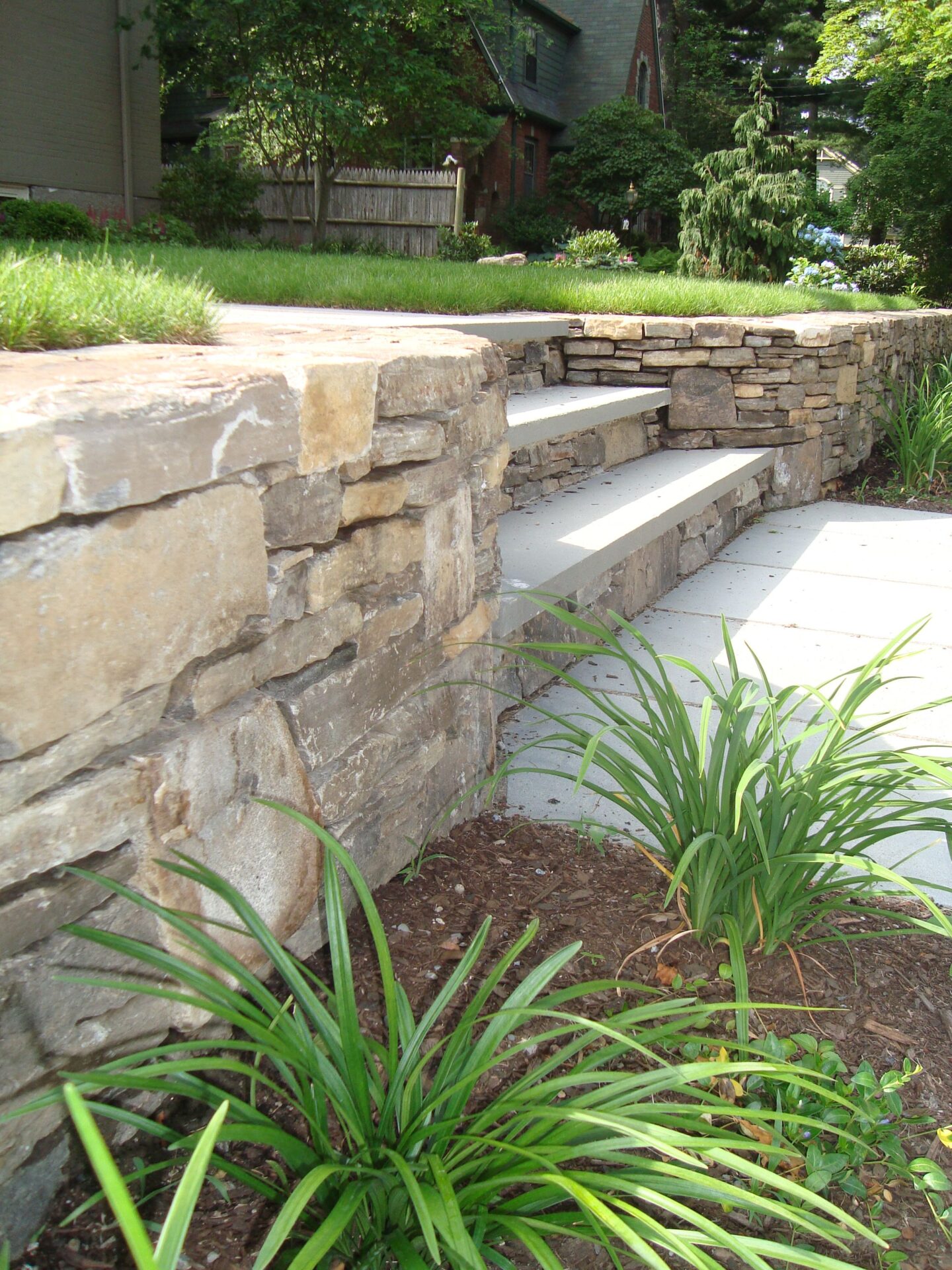 A stone retaining wall with steps, surrounded by green plants, leads to a brick house with a gabled roof and lush garden.
