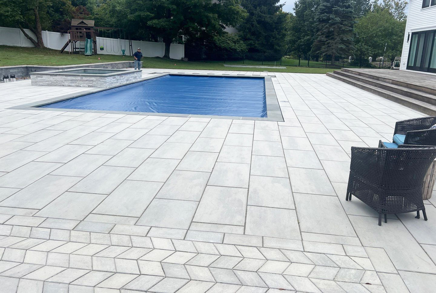 A modern outdoor pool with safety cover, tile patio, two chairs, playground, and trees. A person stands near the pool's edge.