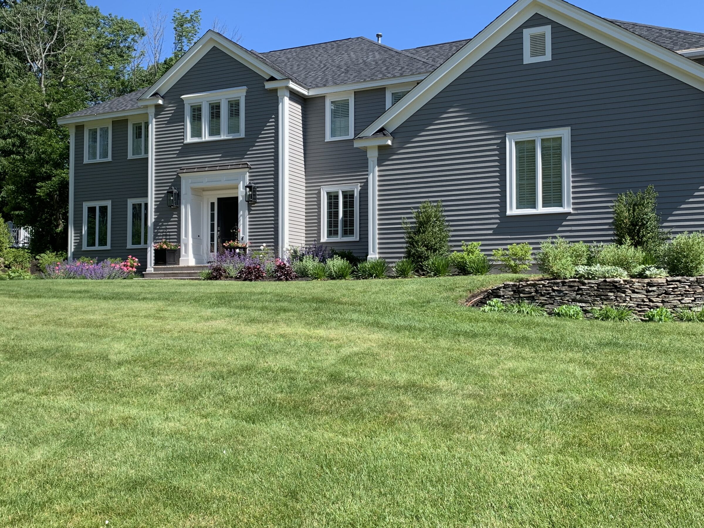 A gray two-story house with a white entrance is surrounded by a well-maintained garden and lush green lawn under clear skies.