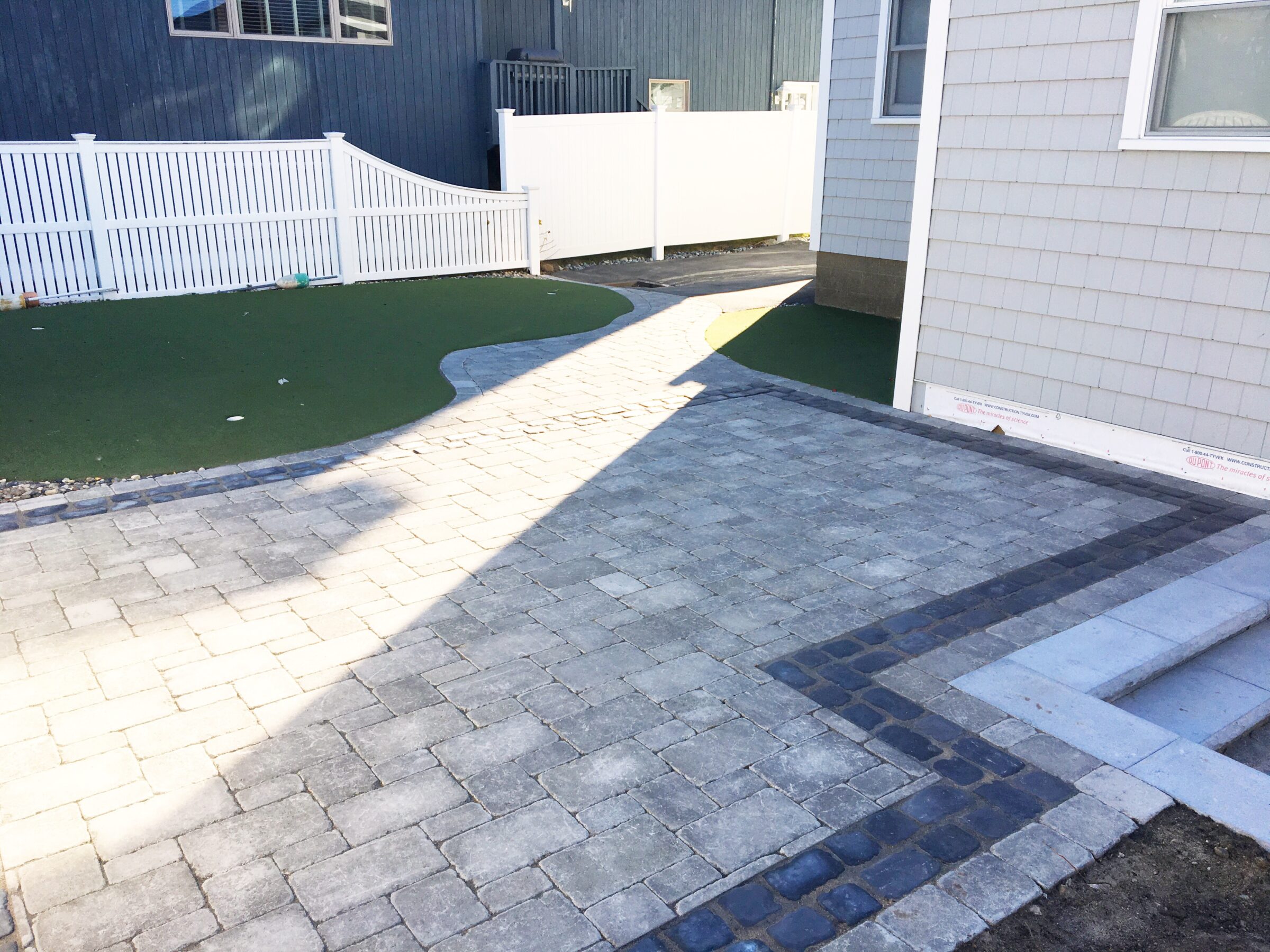 A paved stone path leads between buildings with a white picket fence surrounding a small artificial grass area, under clear sunlight.
