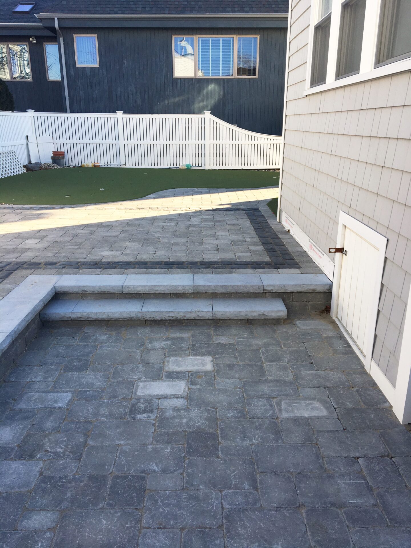 A residential backyard with a stone patio, small steps, white fence, and dark house. Neatly maintained with no visible people or vegetation.