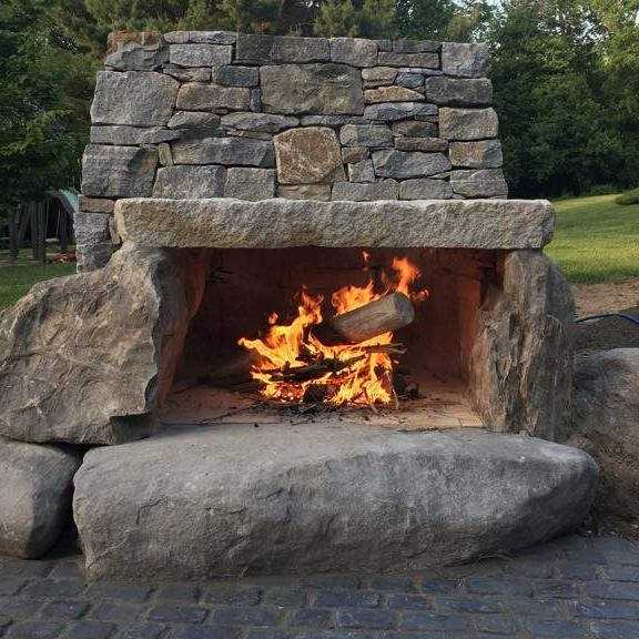 An outdoor stone fireplace with a lively fire, set amidst a grassy area surrounded by trees, looks inviting for gatherings.