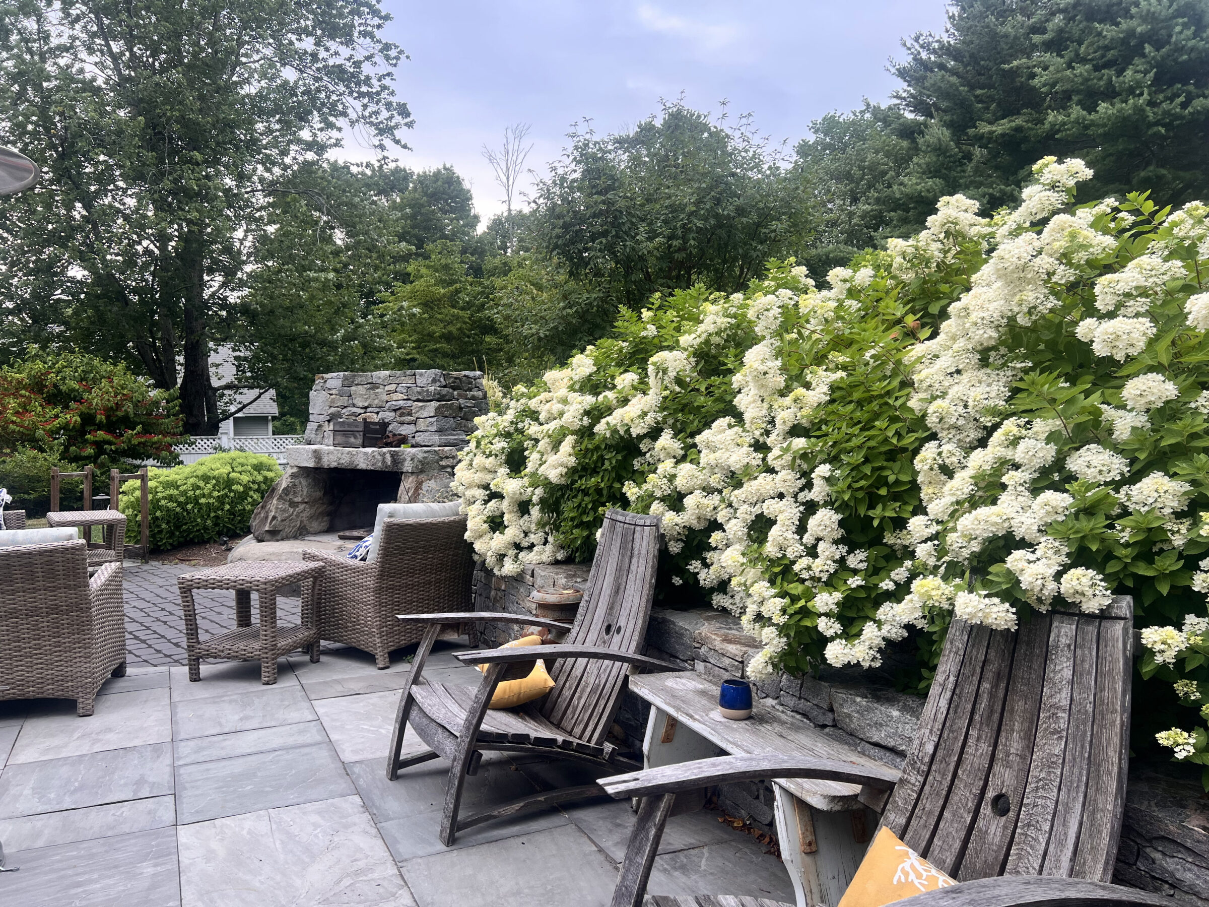 A serene patio with wooden chairs, wicker furniture, lush hydrangea blooms, and a stone fireplace surrounded by trees. Peaceful outdoor setting.