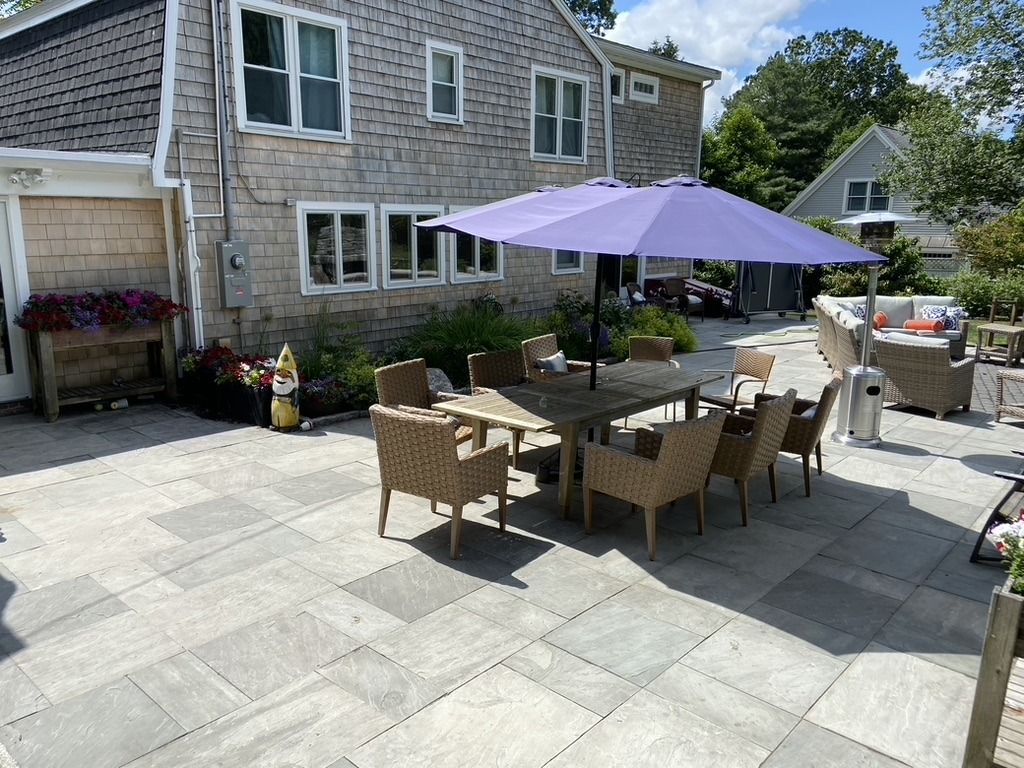 A patio with a dining table, chairs, and a purple umbrella. House exterior features shingles, surrounded by greenery. A gnome decorates the space.