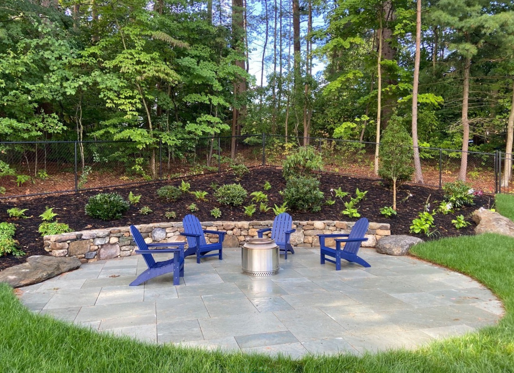 A circular patio with blue chairs and a fire pit, surrounded by greenery and a stone wall, set against a forest backdrop.