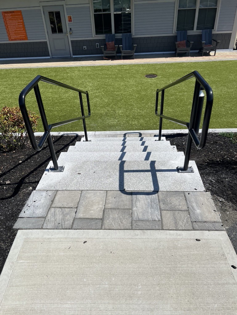A small staircase descends to a paved patio with outdoor seating. There's a building entrance and artificial grass visible in the background.