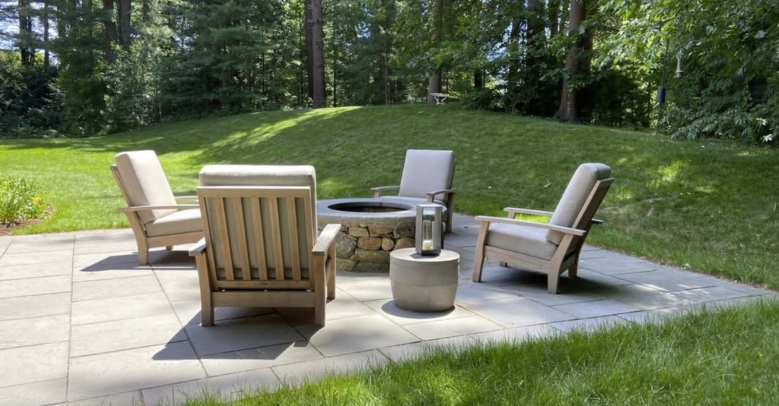A patio with four chairs surrounds a stone fire pit, set against a backdrop of lush, green trees and grass.
