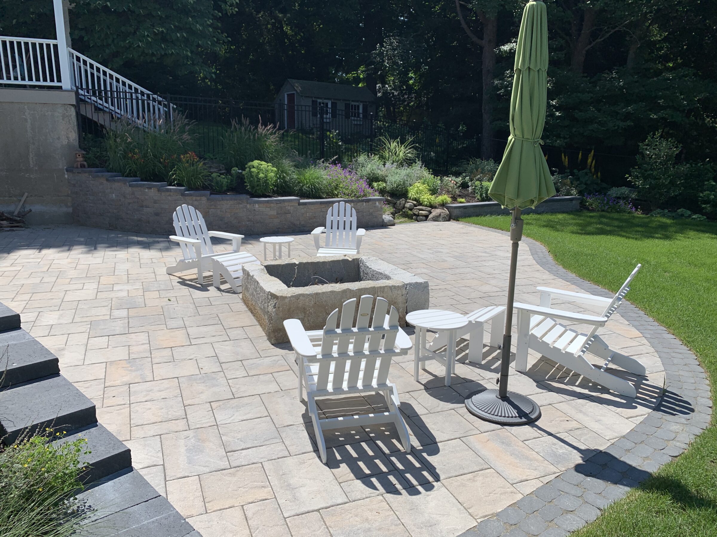 A serene patio with white chairs encircles a stone fire pit. A closed green umbrella stands nearby, surrounded by lush greenery.