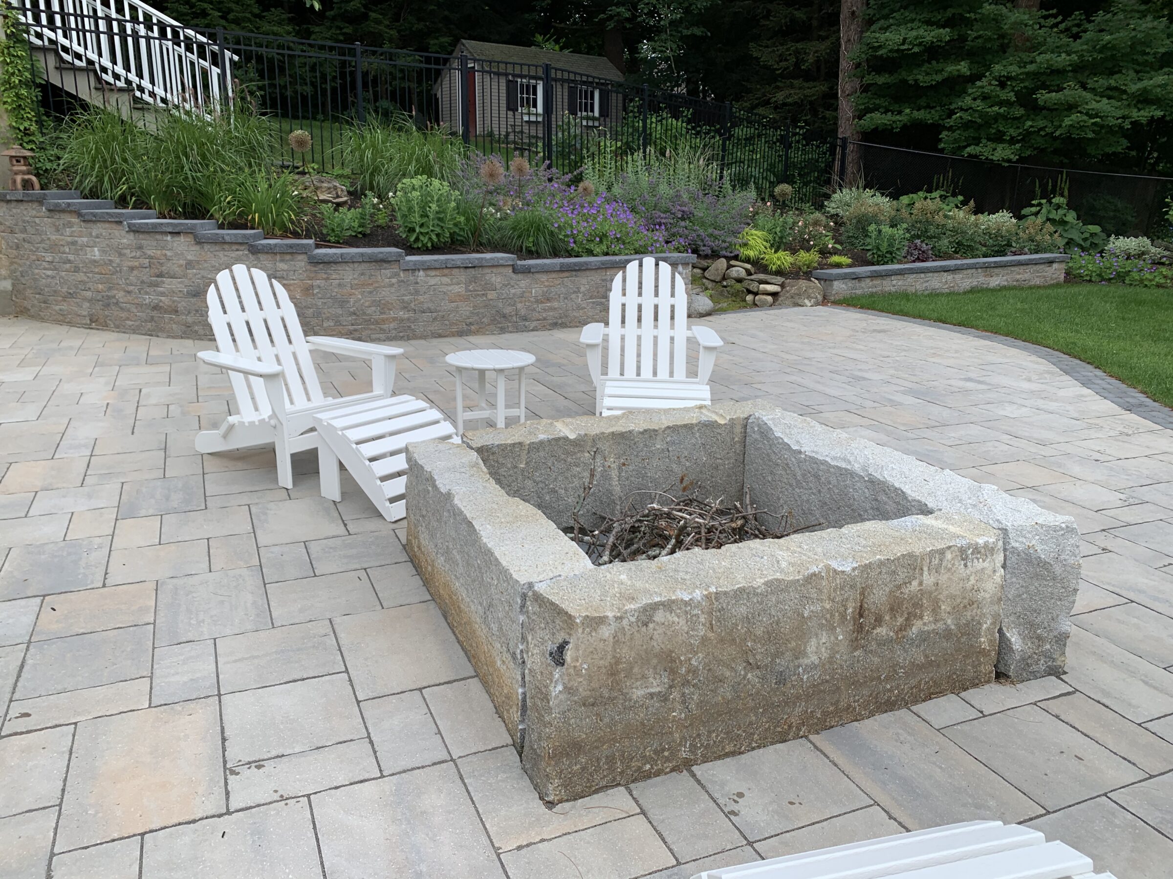 Patio with white Adirondack chairs, stone fire pit, and landscaped garden with flowers and greenery. Peaceful outdoor setting, no people present.