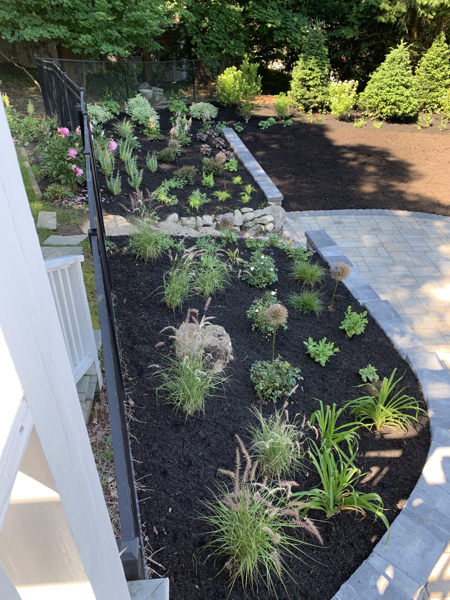 A landscaped garden with various plants and flowers, surrounded by trees, features a curved pathway with a stone border and black mulch.