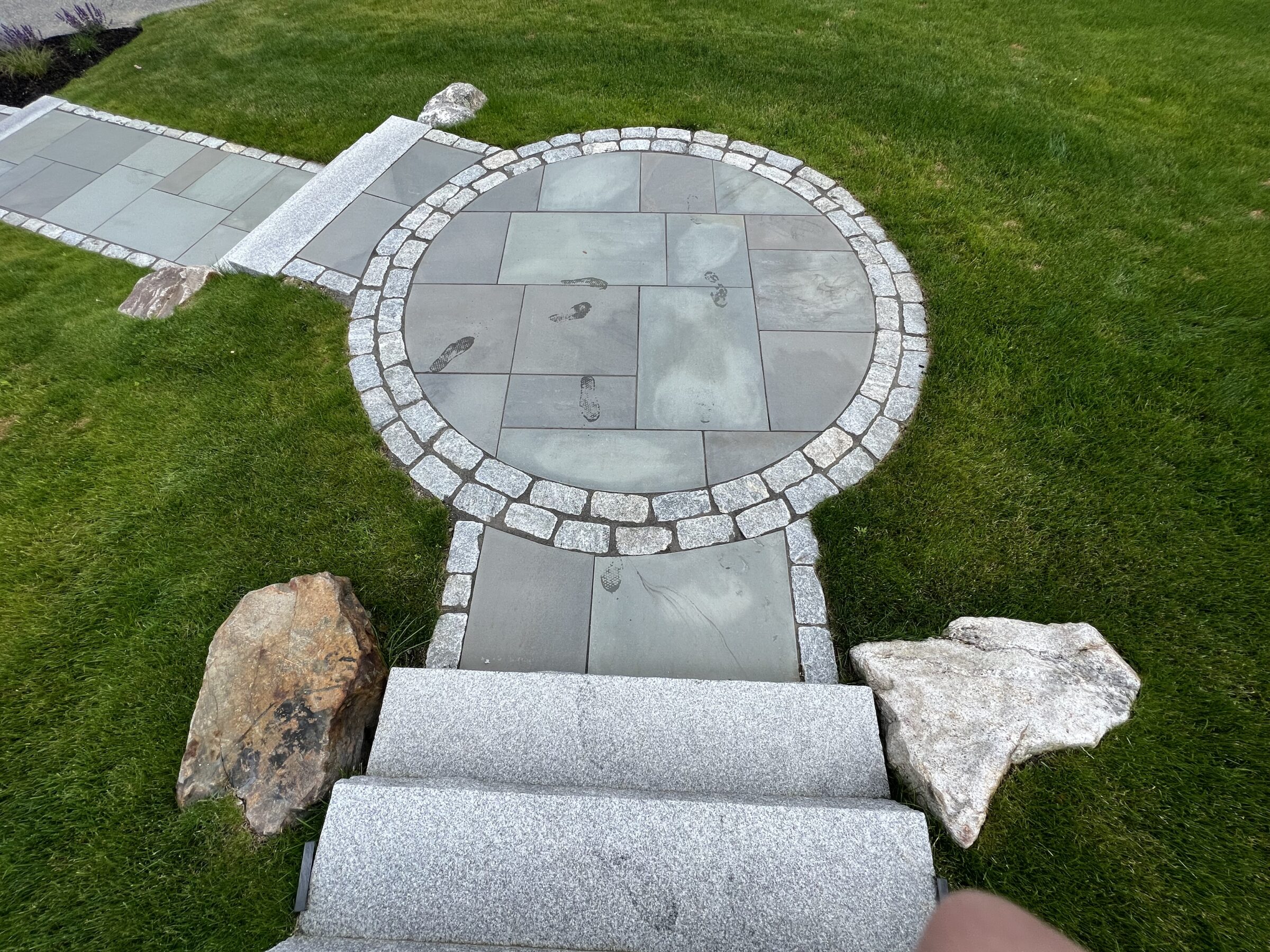 A circular paved stone area with visible footprints leads to a pathway, surrounded by grass and bordered by large rocks.