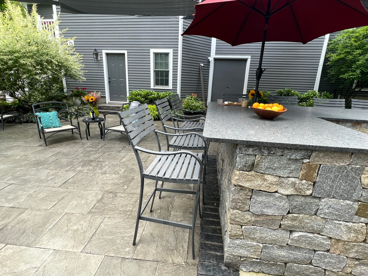 Outdoor patio with stone bar, red umbrella, and seating. Bowl of oranges on counter. Gray siding house and green plants surround.