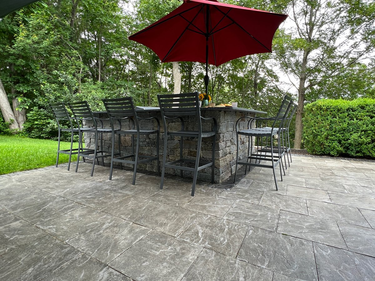 A stone bar with high chairs and a red umbrella stands on a patio surrounded by lush green trees and bushes.