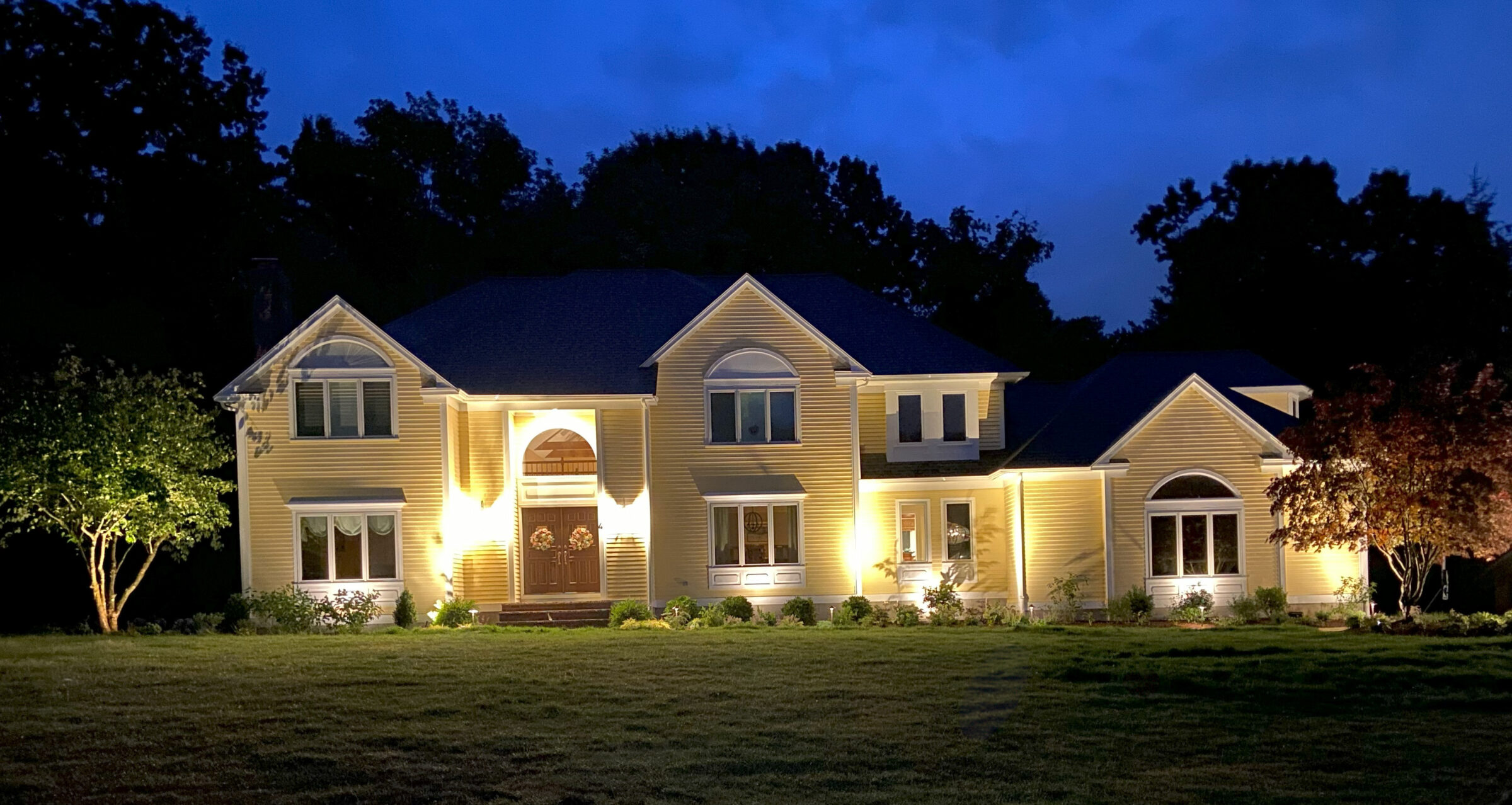 A well-lit, large yellow house with a dark roof is surrounded by trees at dusk, creating a serene and inviting atmosphere.