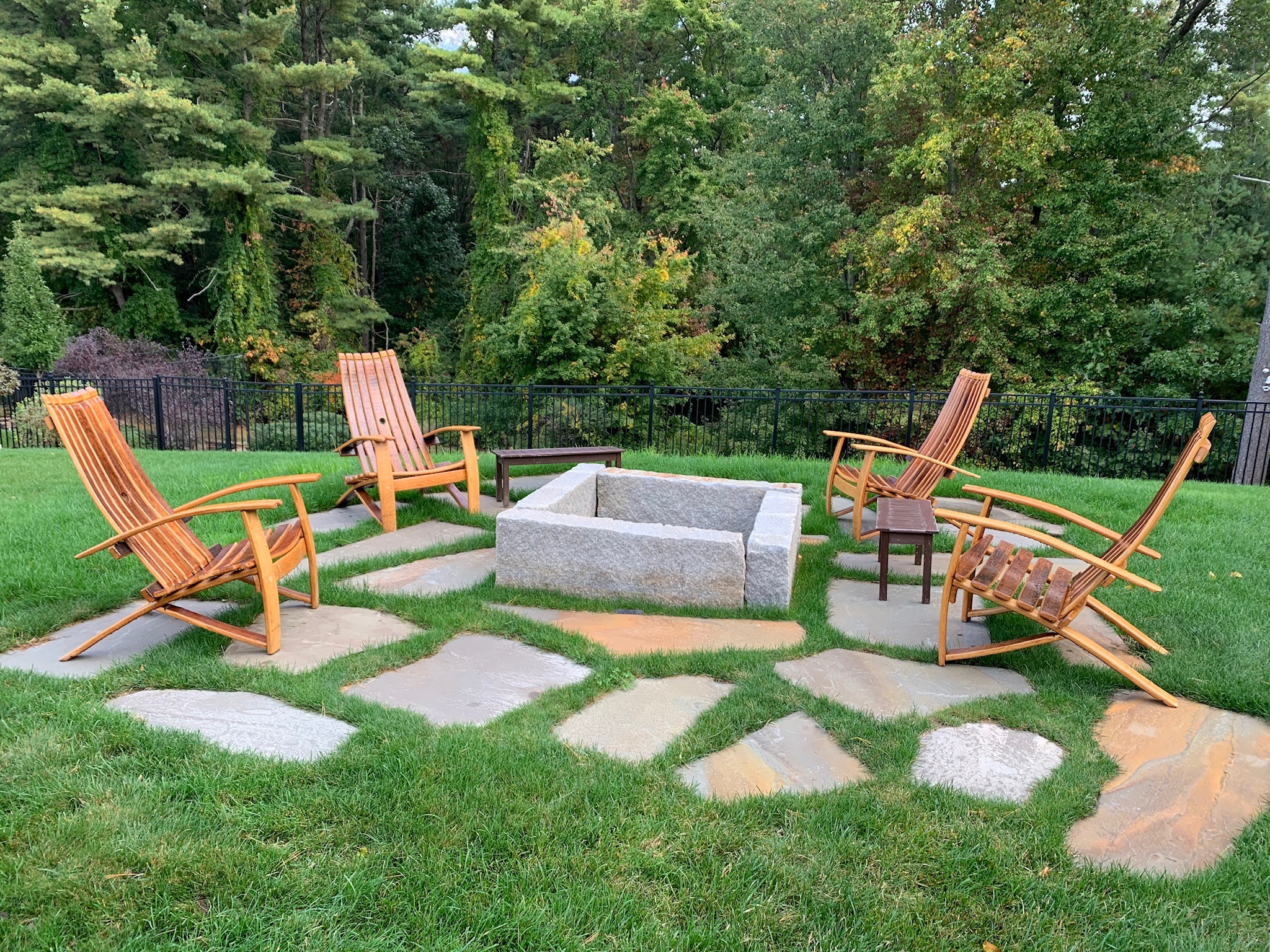Four wooden chairs encircle a square fire pit on a grassy lawn, surrounded by trees and stone paving. Relaxing outdoor setting.