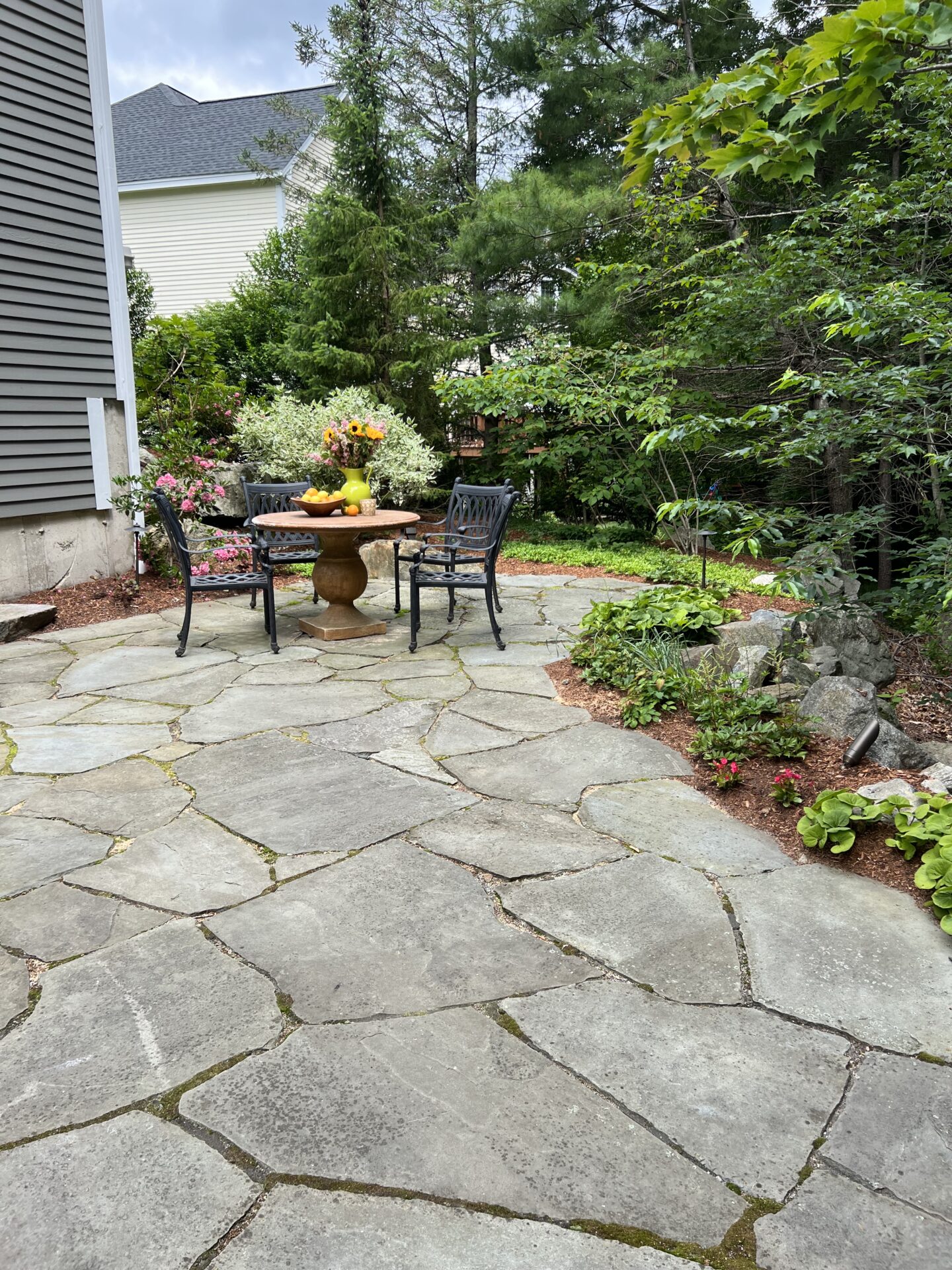 Stone patio with round table and chairs surrounded by lush greenery and flowers, adjacent to a residential house with gray siding.