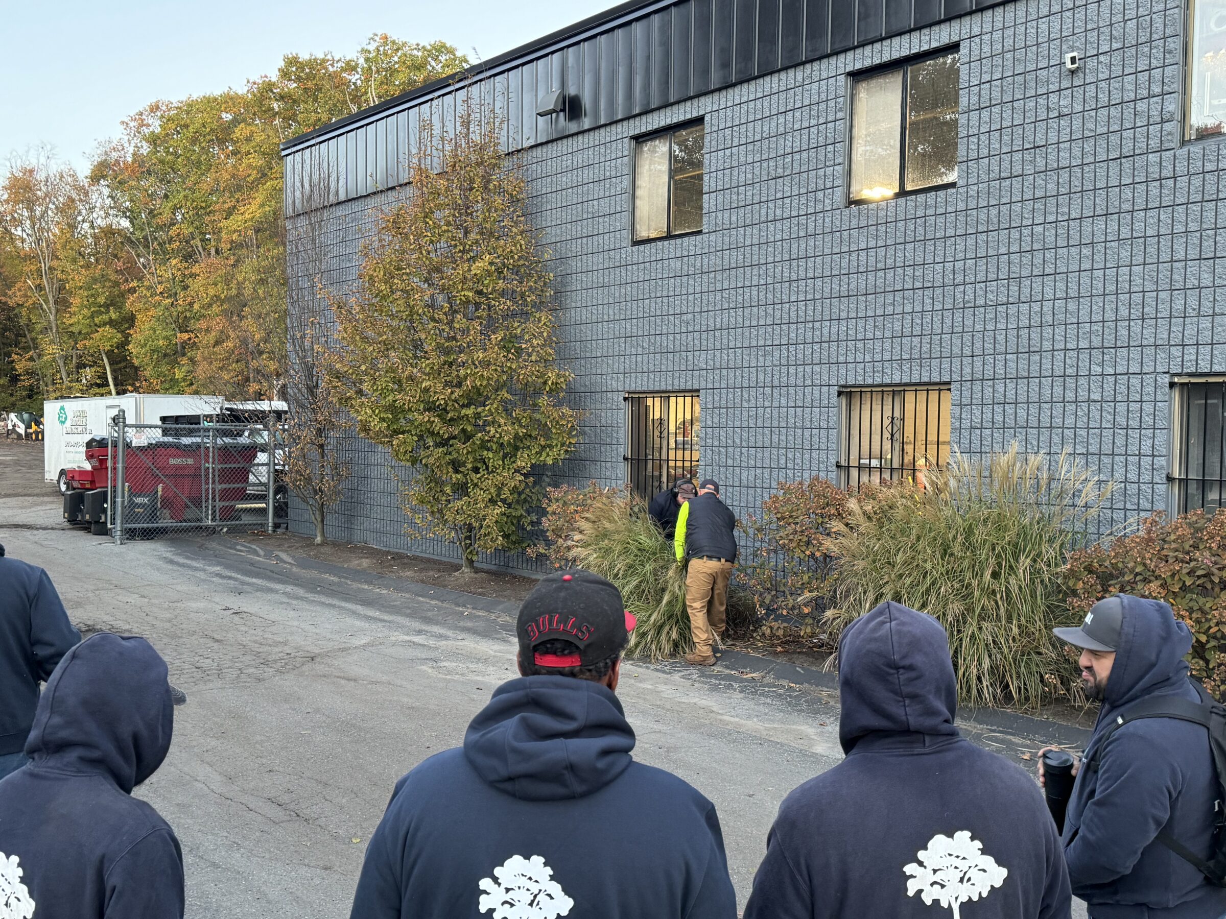 A group of people wearing hoodies stand near a gray building, observing two others working by windows, with autumn trees nearby.