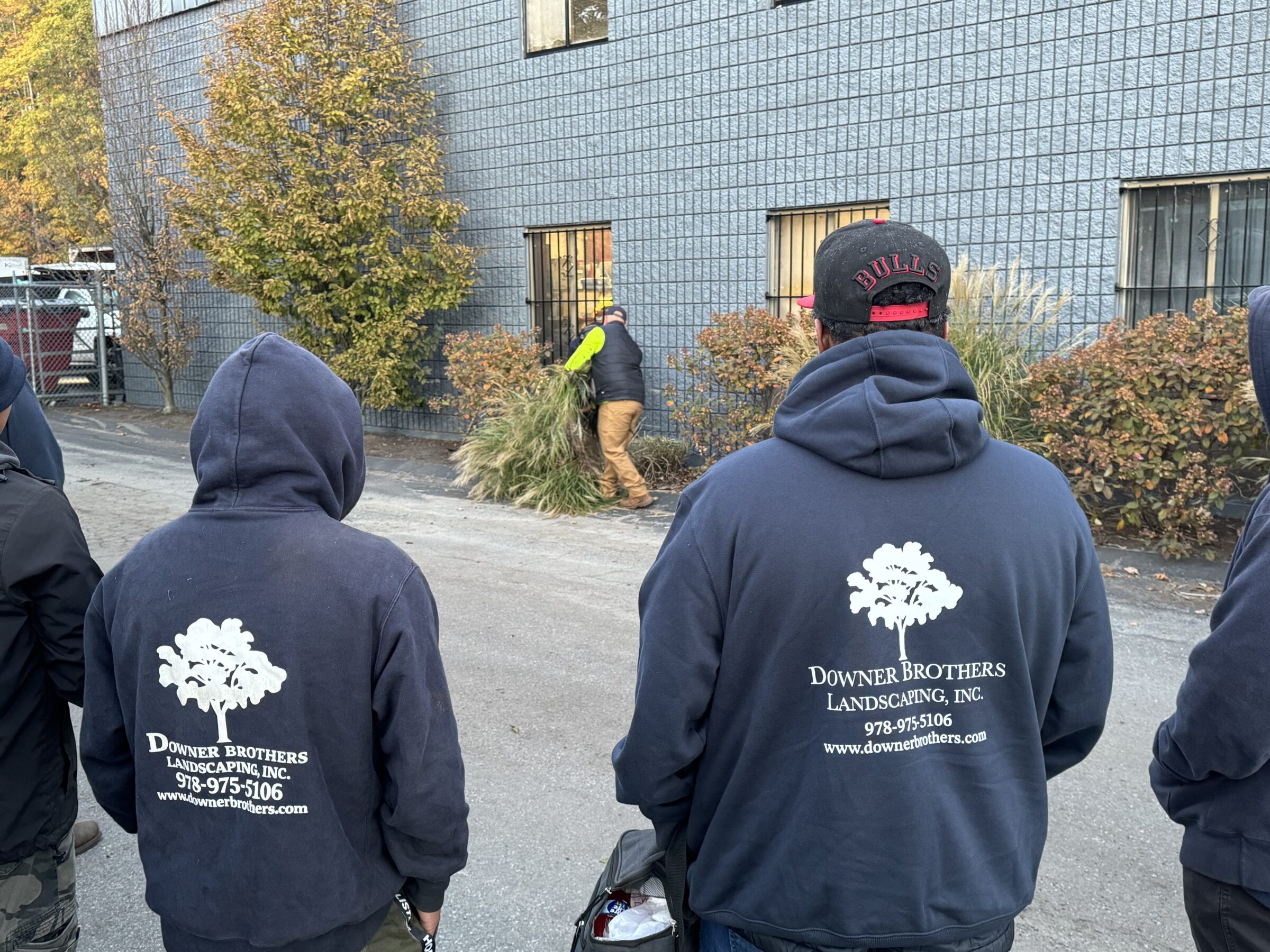 A group of people in hooded sweatshirts, with "Downer Brothers Landscaping, Inc." logo, watch a person tending to plants by a building.