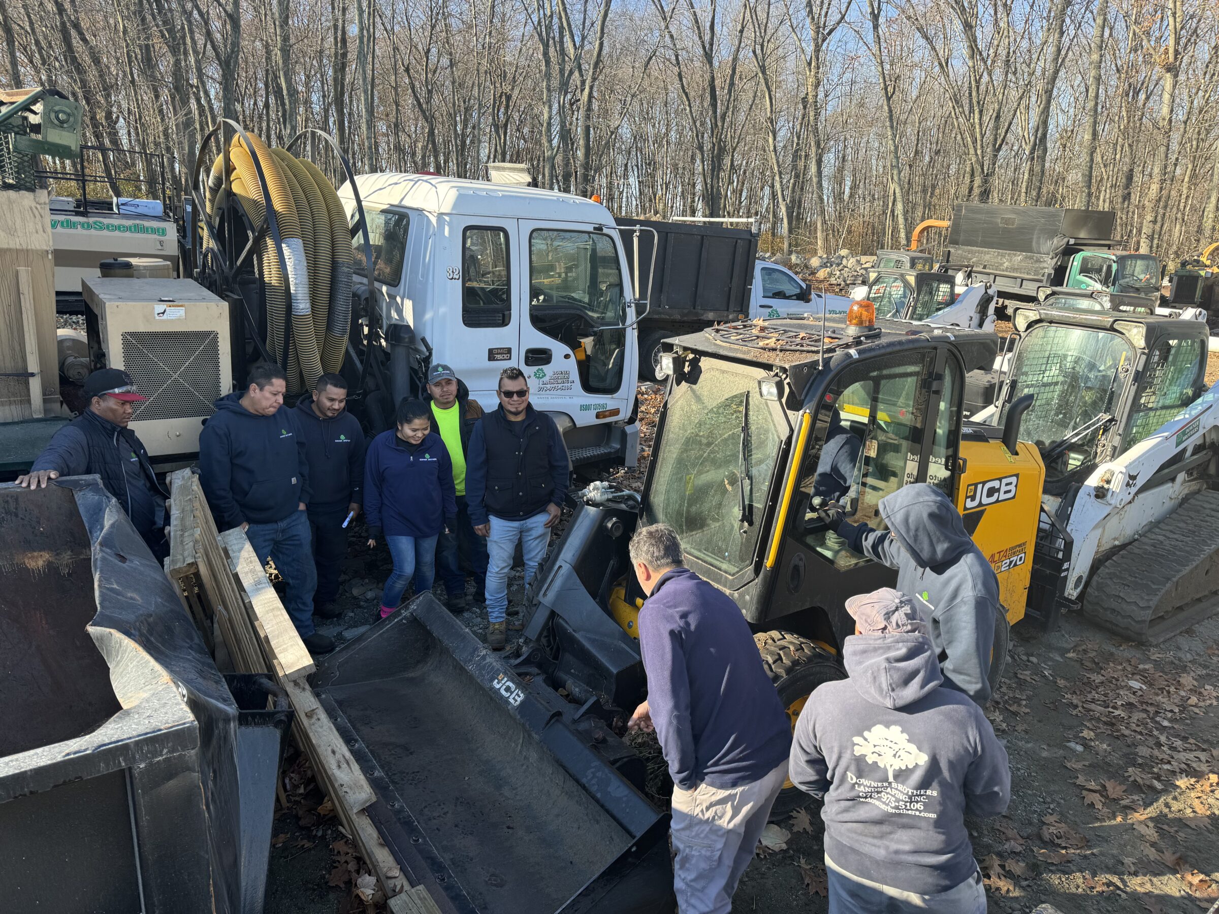 A group of people gather around construction vehicles in a wooded area, examining equipment. Various trucks and machinery are visible in the background.