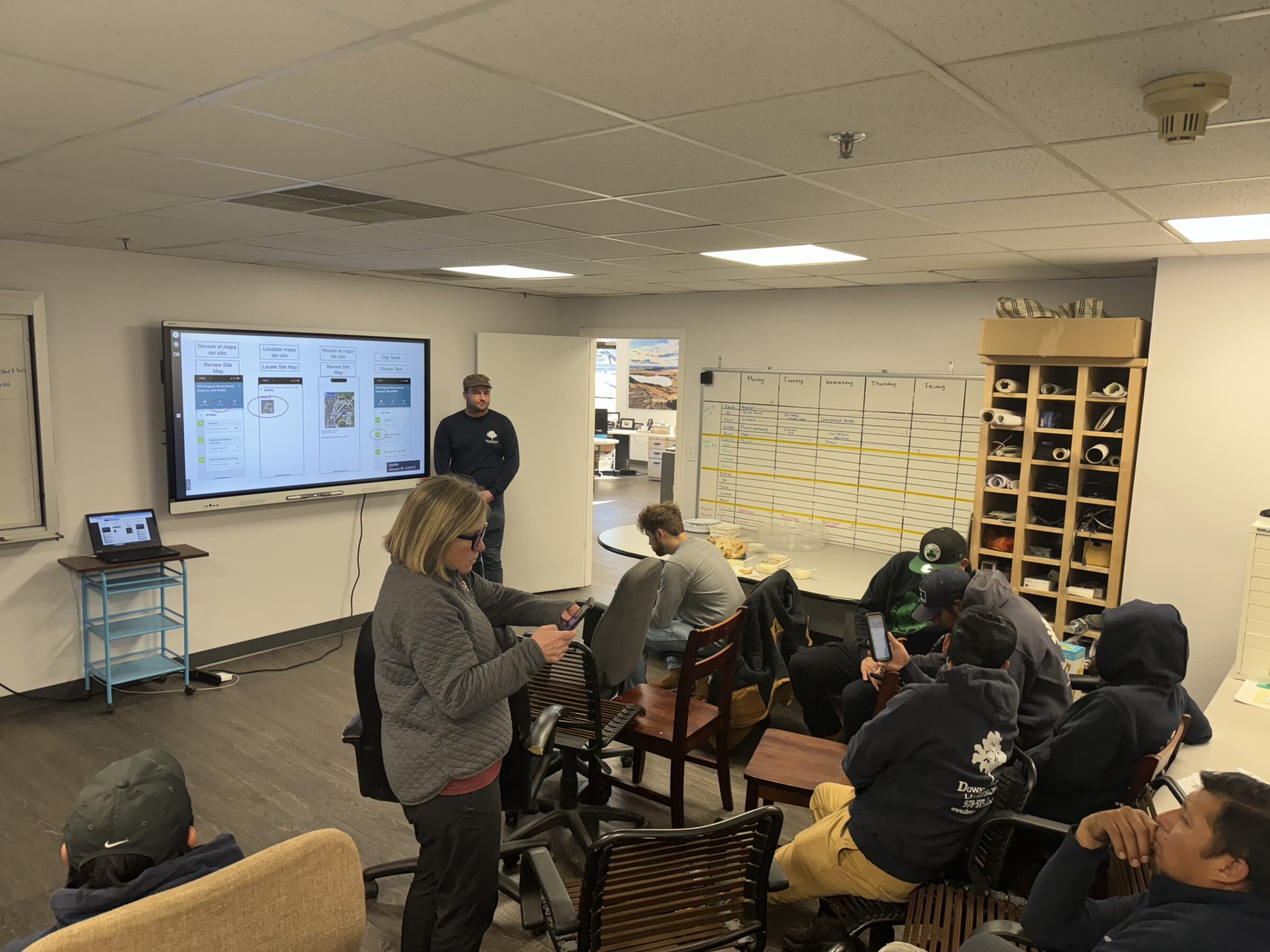 A classroom setting features a presentation, with people seated and one standing. A digital screen and whiteboard with schedules are visible.