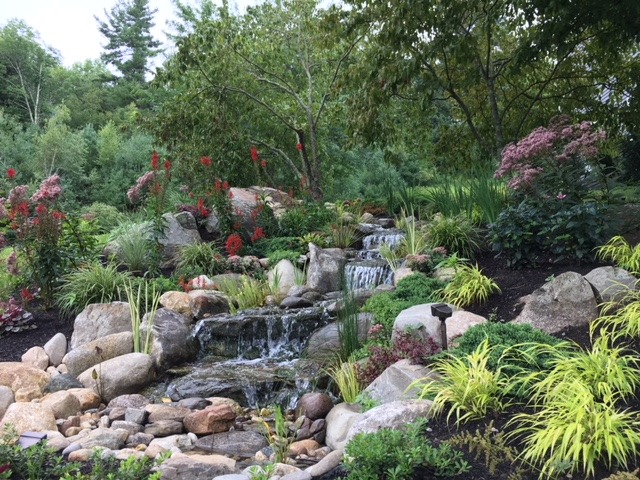 A serene garden with cascading waterfalls, vibrant flowers, lush greenery, and rocks, surrounded by trees under a clear sky, creating a tranquil scene.