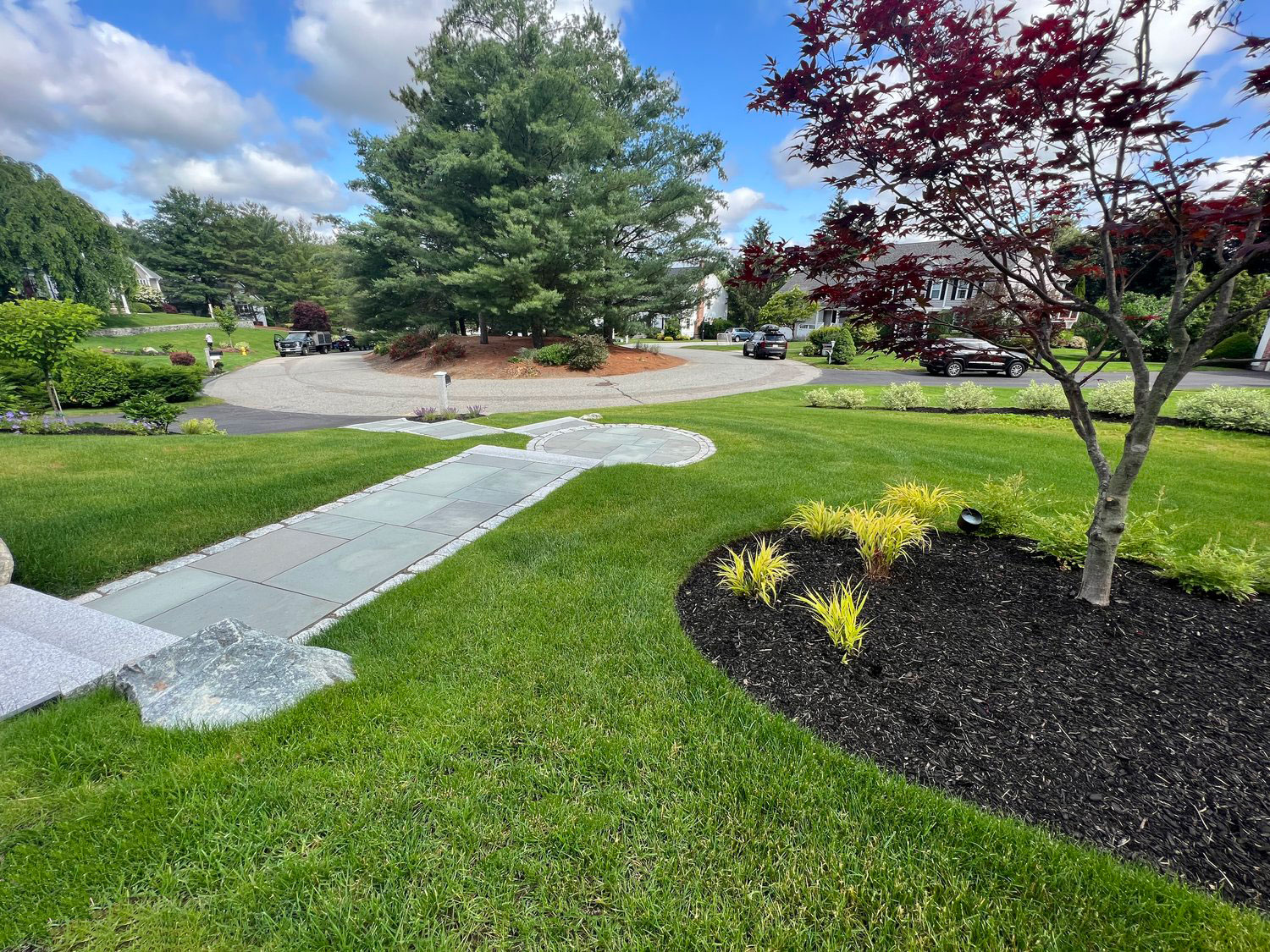A landscaped suburban neighborhood with a curved driveway, green lawn, and trees under a partly cloudy sky. No recognizable landmarks or historical buildings.