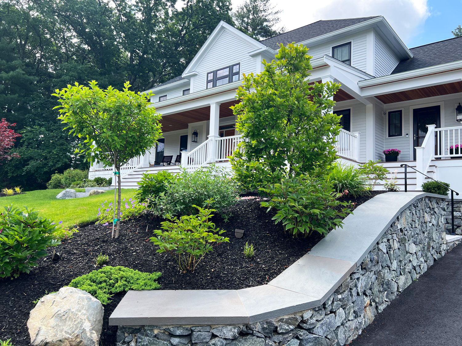 A beautiful white house with a porch, surrounded by vibrant greenery and a well-maintained stone wall in a lush garden setting.