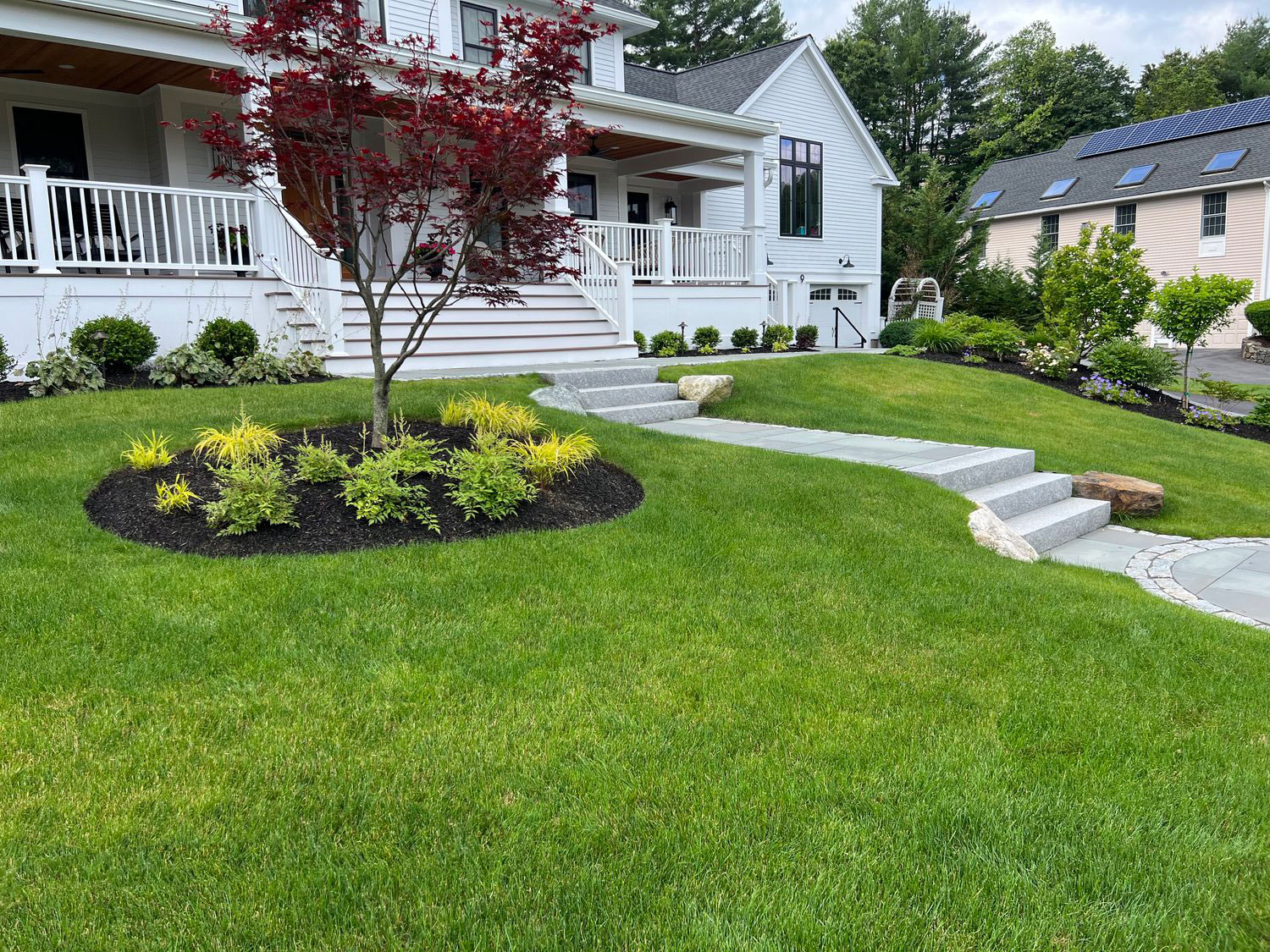 A well-maintained lawn with landscaping leads to a house featuring white railings, steps, and a porch, surrounded by lush greenery and trees.