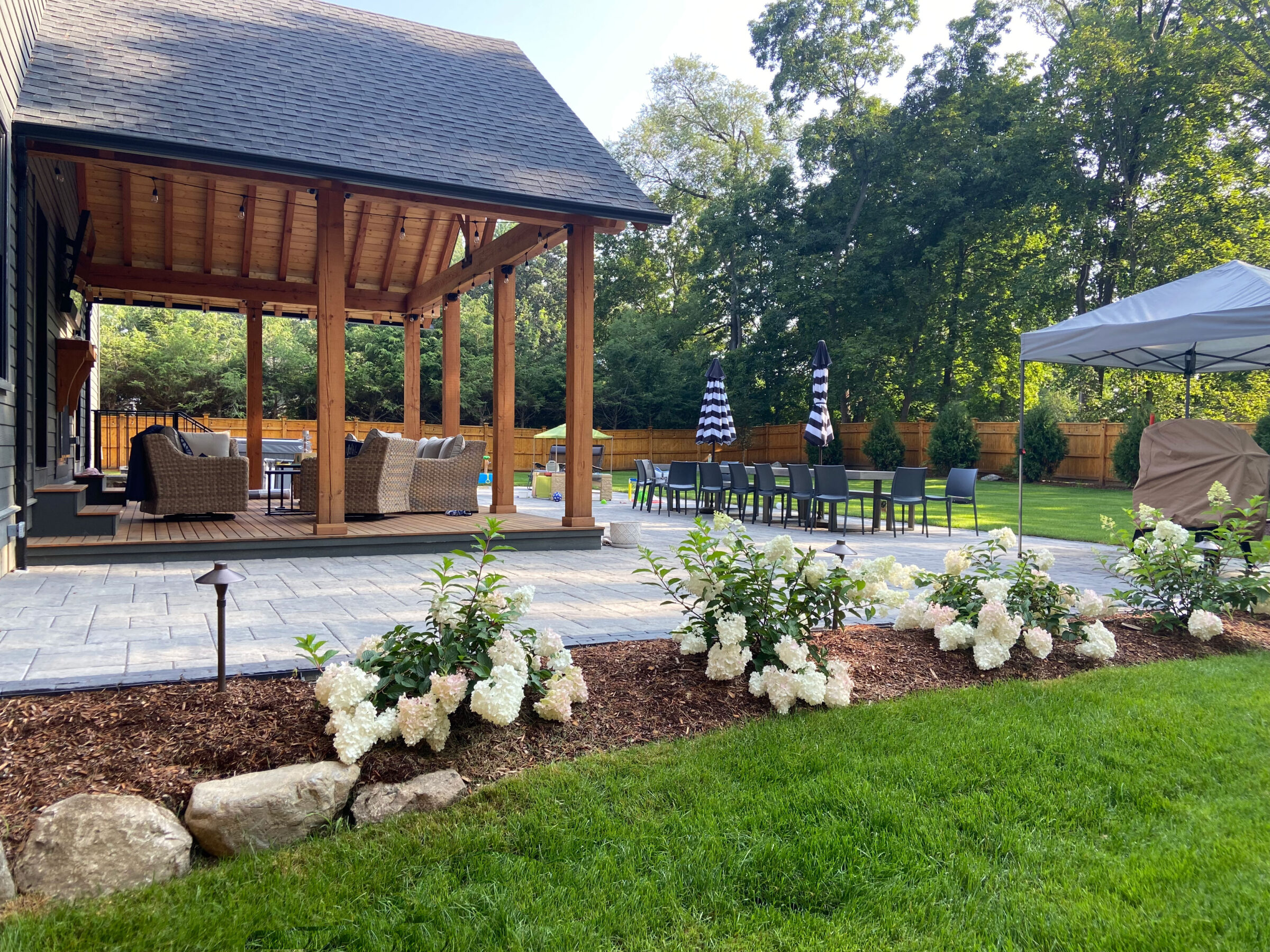 A backyard patio features wicker furniture, a dining area with striped umbrellas, and beautiful hydrangeas, set against a lush, tree-lined backdrop.