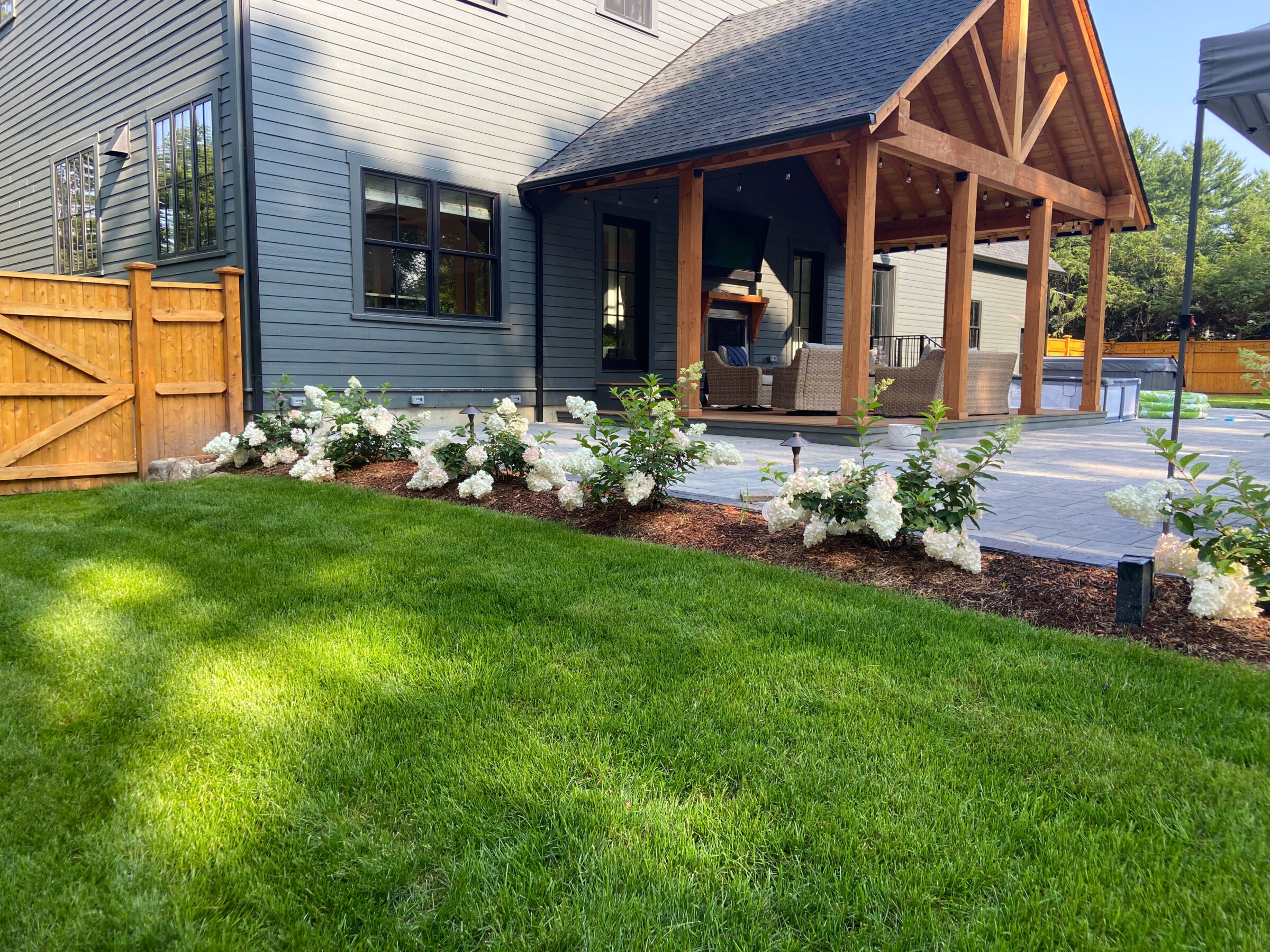 A landscaped backyard features a modern house with a wooden porch, neatly arranged flower beds, and a green lawn under a clear sky.