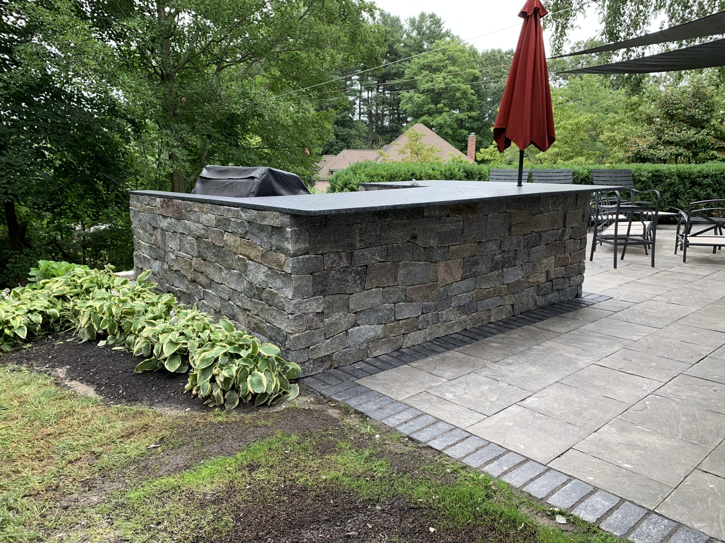 A stone outdoor kitchen with countertop, a covered grill, red umbrella, chairs, and lush greenery, set in a serene, landscaped garden.