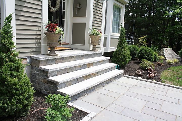 A residential entrance features stone steps with potted plants, surrounded by landscaped greenery and shrubs, leading to a house with siding and large windows.