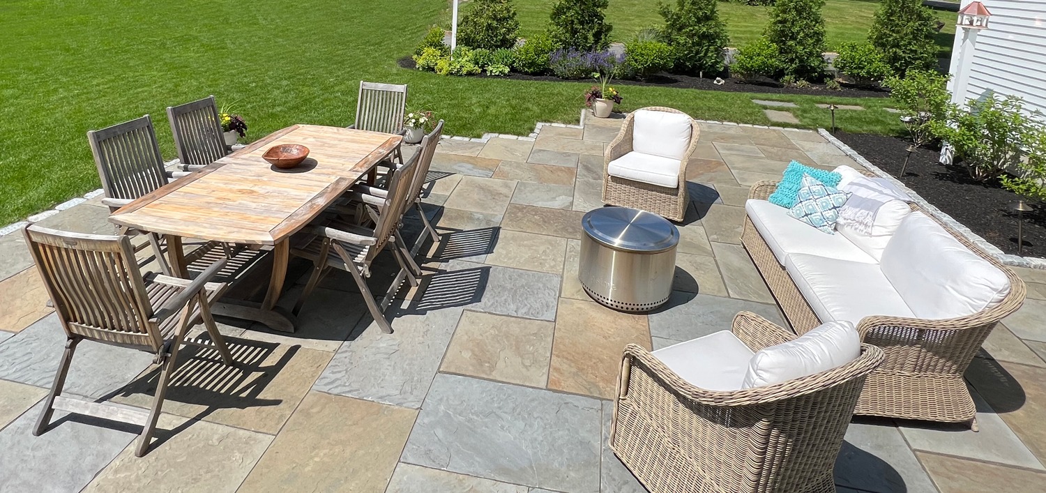 Outdoor patio with wooden dining table, wicker seating, and metal fire pit. Surrounded by a manicured lawn and lush greenery.