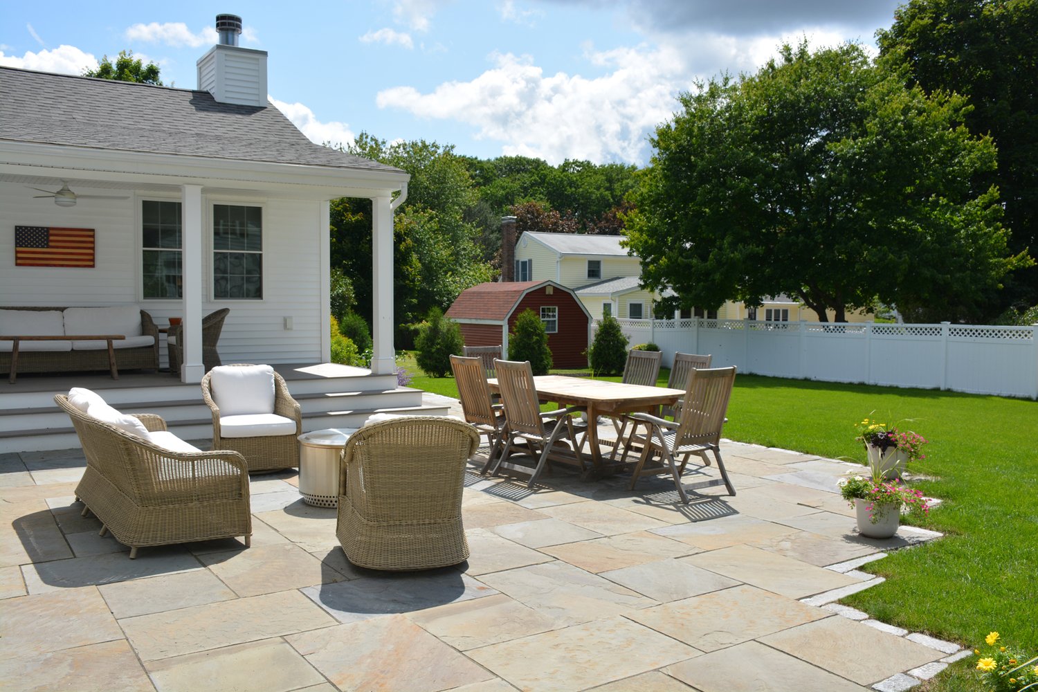A spacious backyard with patio furniture, a white house, and vibrant green lawn, featuring planters and an American flag wall decor.