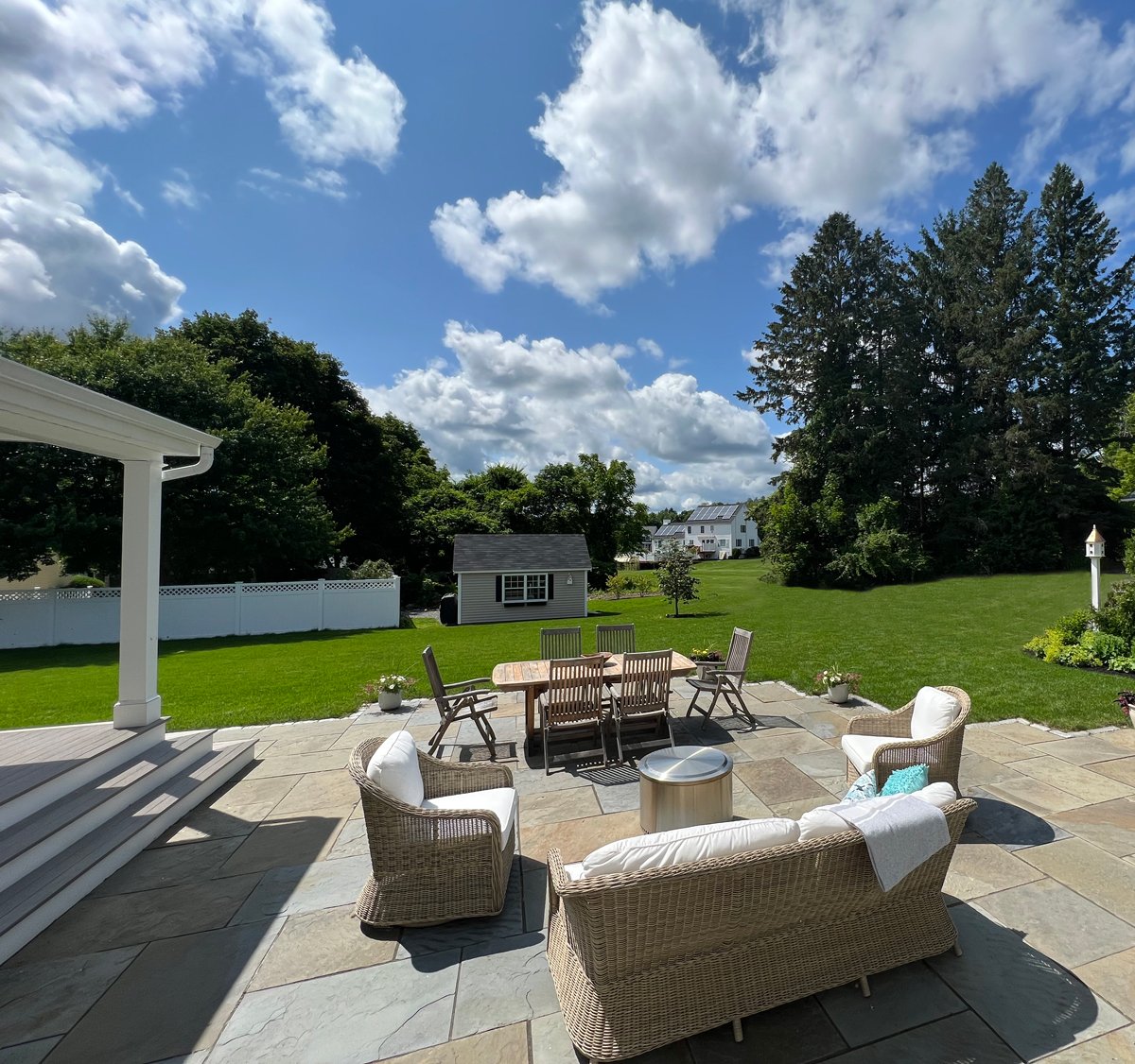 Spacious patio with wicker furniture overlooks lush backyard, trees, and a small shed under a blue sky with scattered clouds.