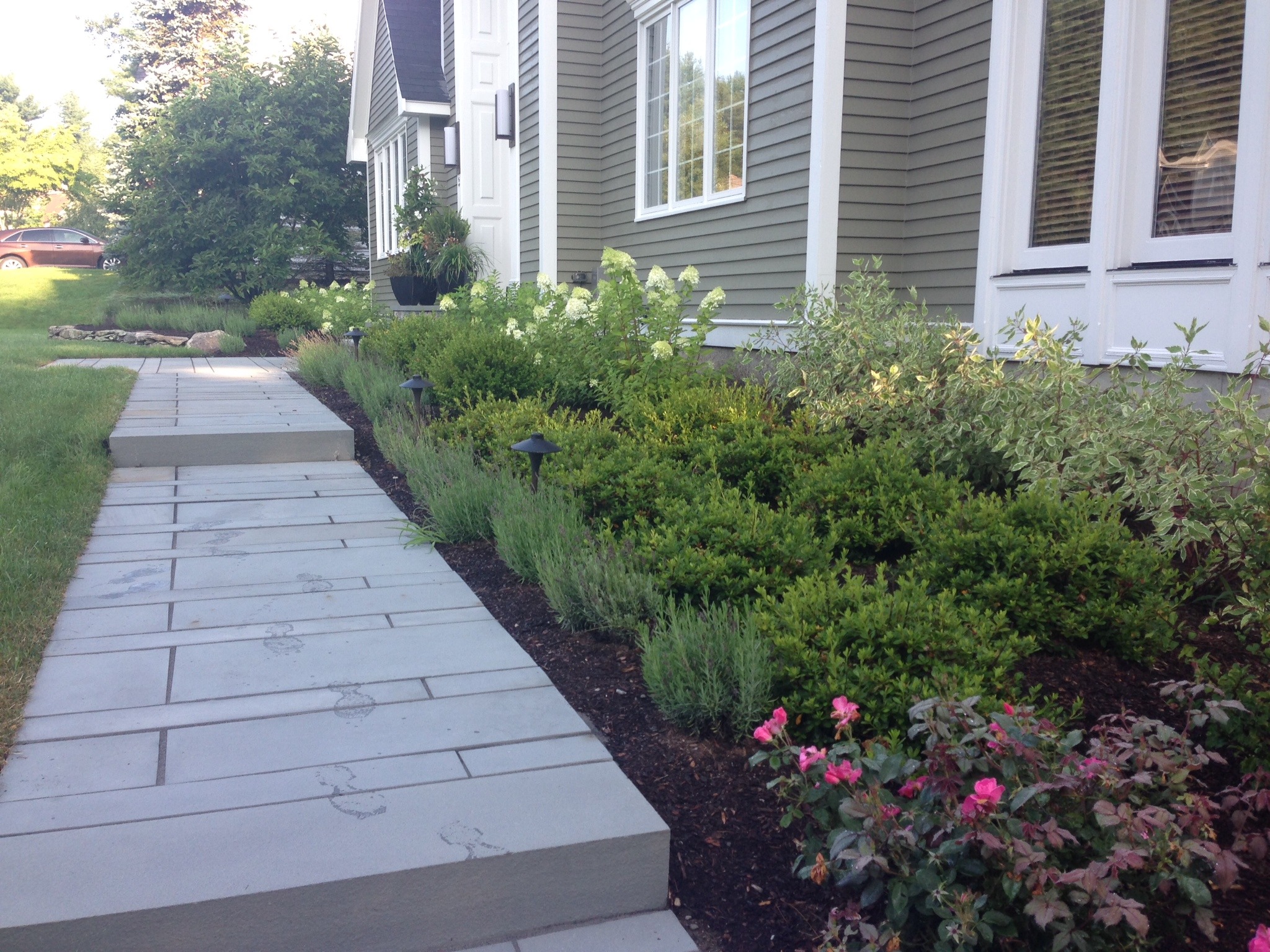 A modern house with a green facade, surrounded by neatly trimmed shrubs, colorful flowers, and a paved pathway leading to the entrance.