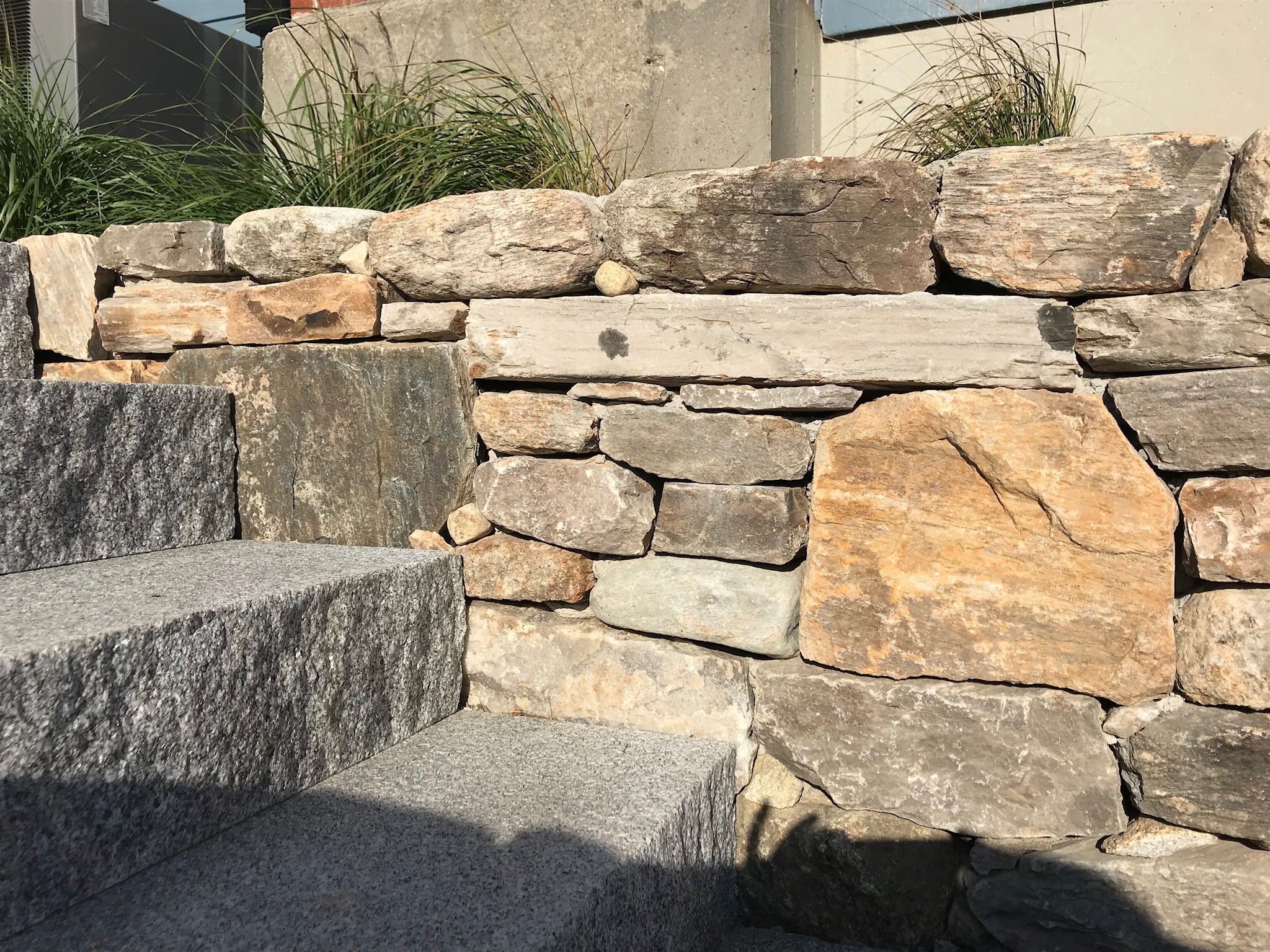 Stone wall and granite steps with sunlight casting shadows, surrounded by green grass. Rough, natural stones create a rustic, textured appearance.