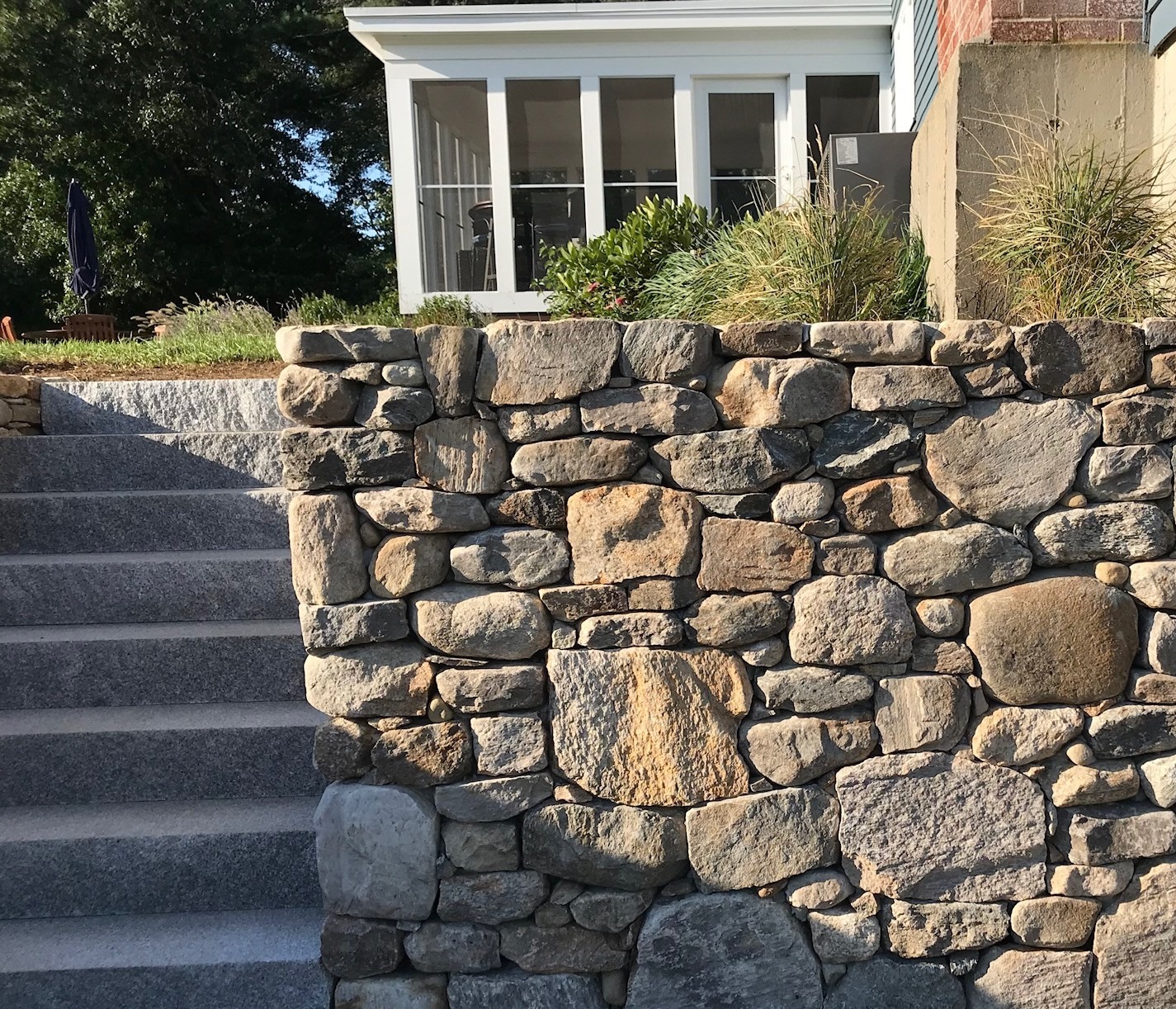 Stone wall with steps leads to a porch surrounded by greenery. Sunlit area with a closed umbrella in the background.