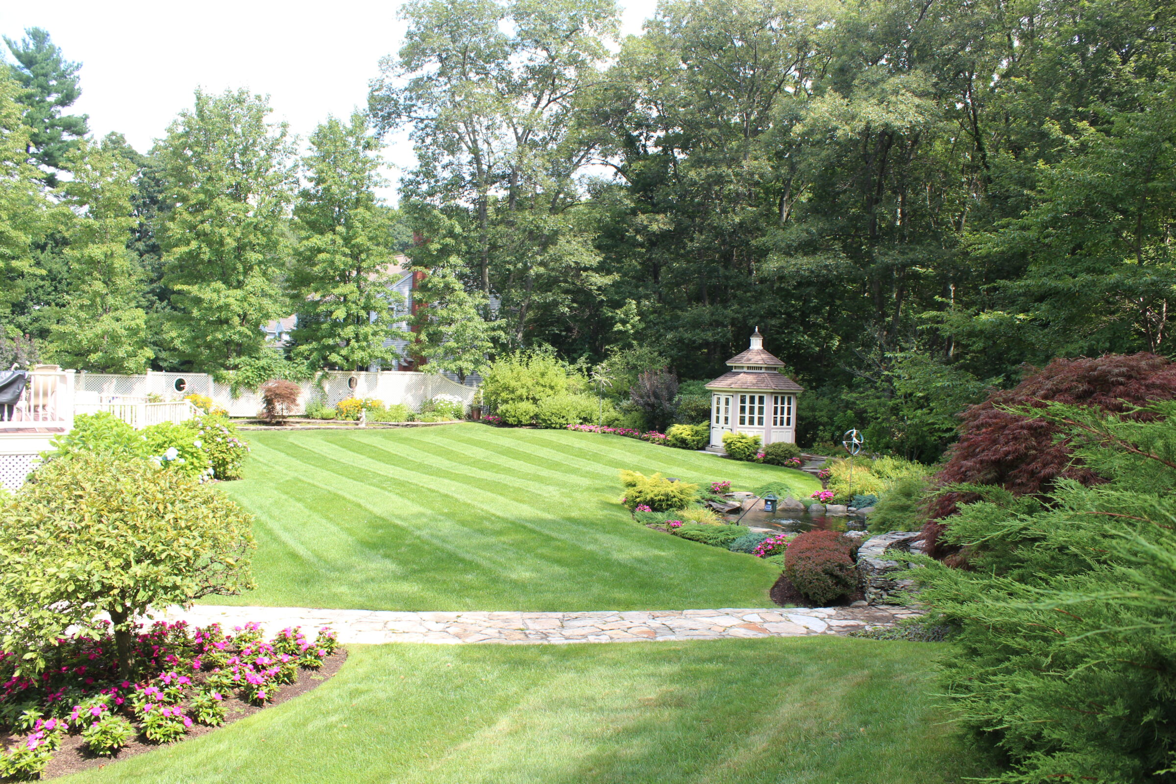 A lush garden with striped lawn, colorful flowers, and a quaint gazebo surrounded by dense trees. Pathways enhance the serene landscape.