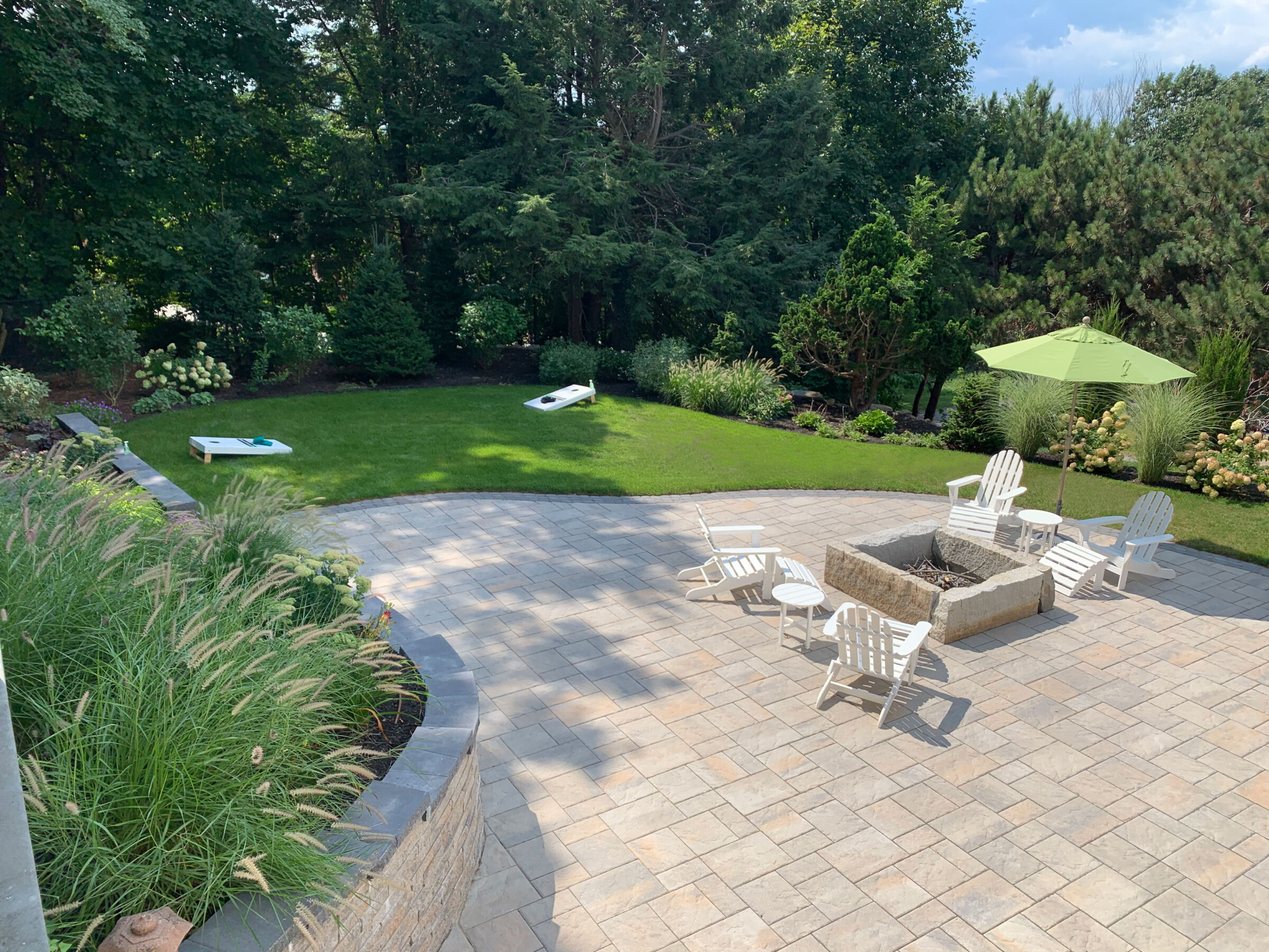 A landscaped backyard with a fire pit, white chairs, green umbrella, and cornhole boards on a grassy area surrounded by trees.